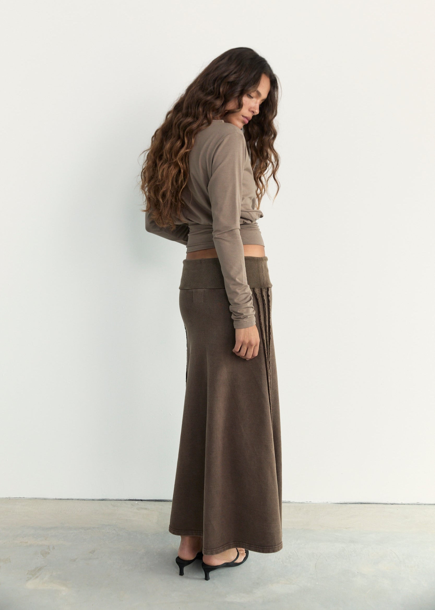 A woman wearing a taupe long-sleeve crop top and a matching brown wide-leg midi skirt, standing in profile against a white wall.