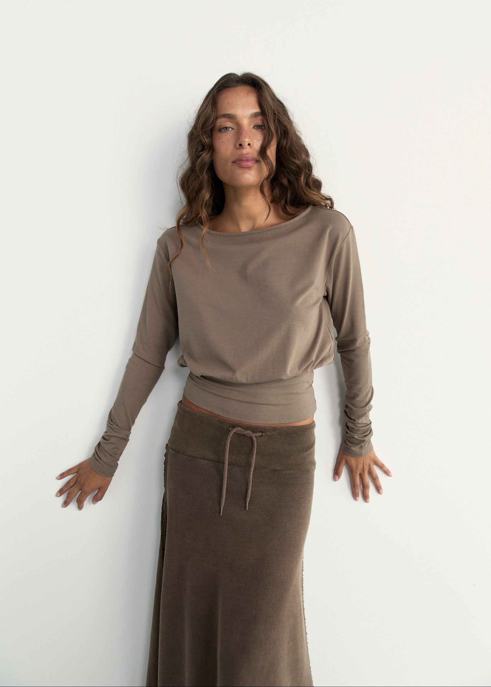 A woman modeling a taupe long-sleeve knit top and a brown skirt against a plain white wall.
