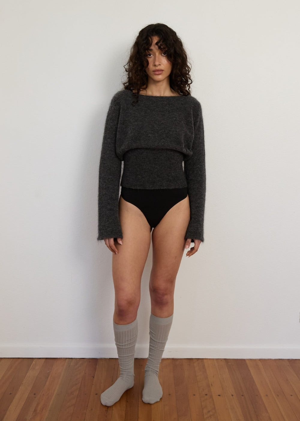Person wearing a dark gray sweater and black underwear against a white wall.
