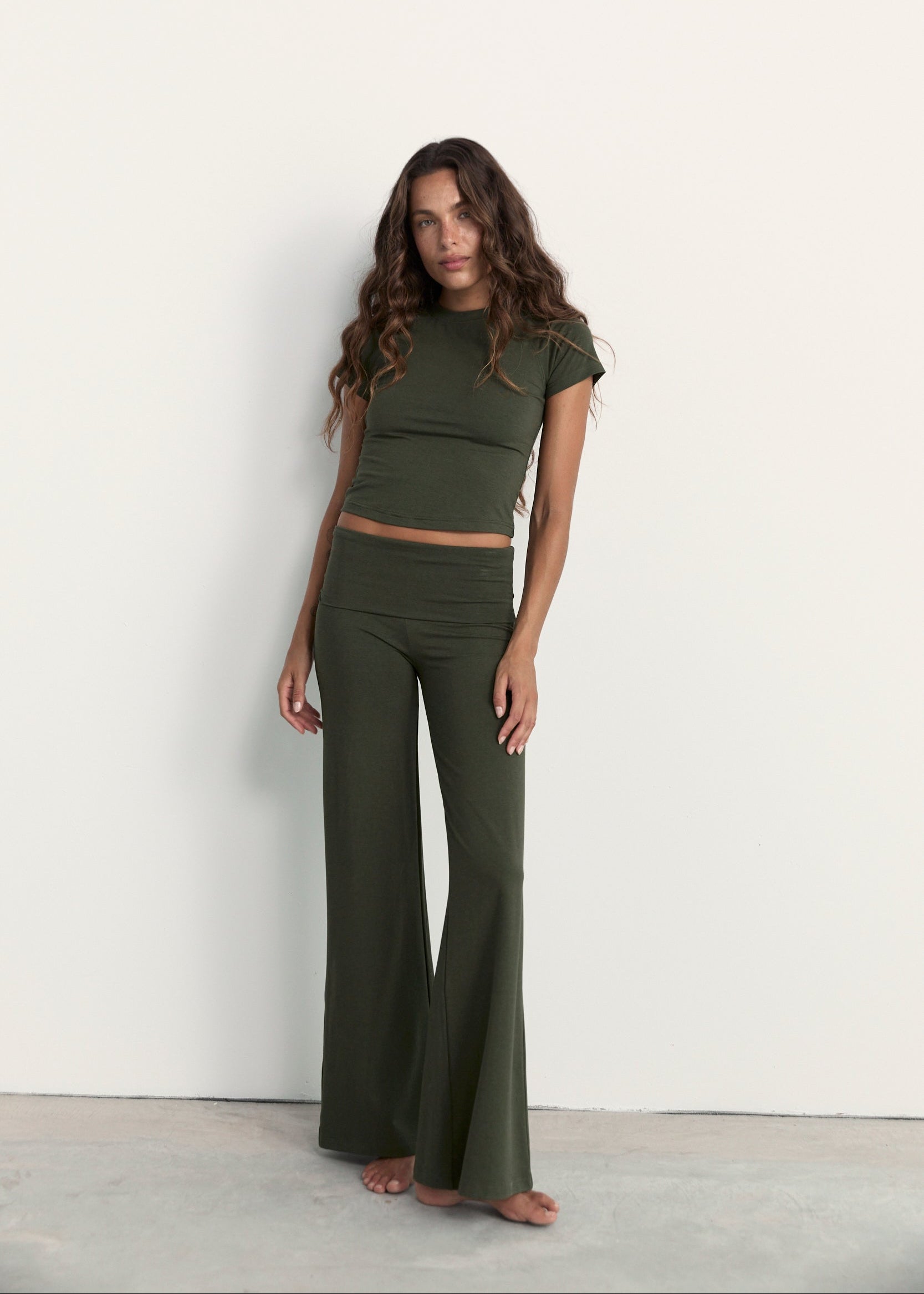 A woman modeling an olive green two-piece lounge set consisting of a short-sleeve crop top and matching high-waisted flared pants, standing against a plain off-white wall.