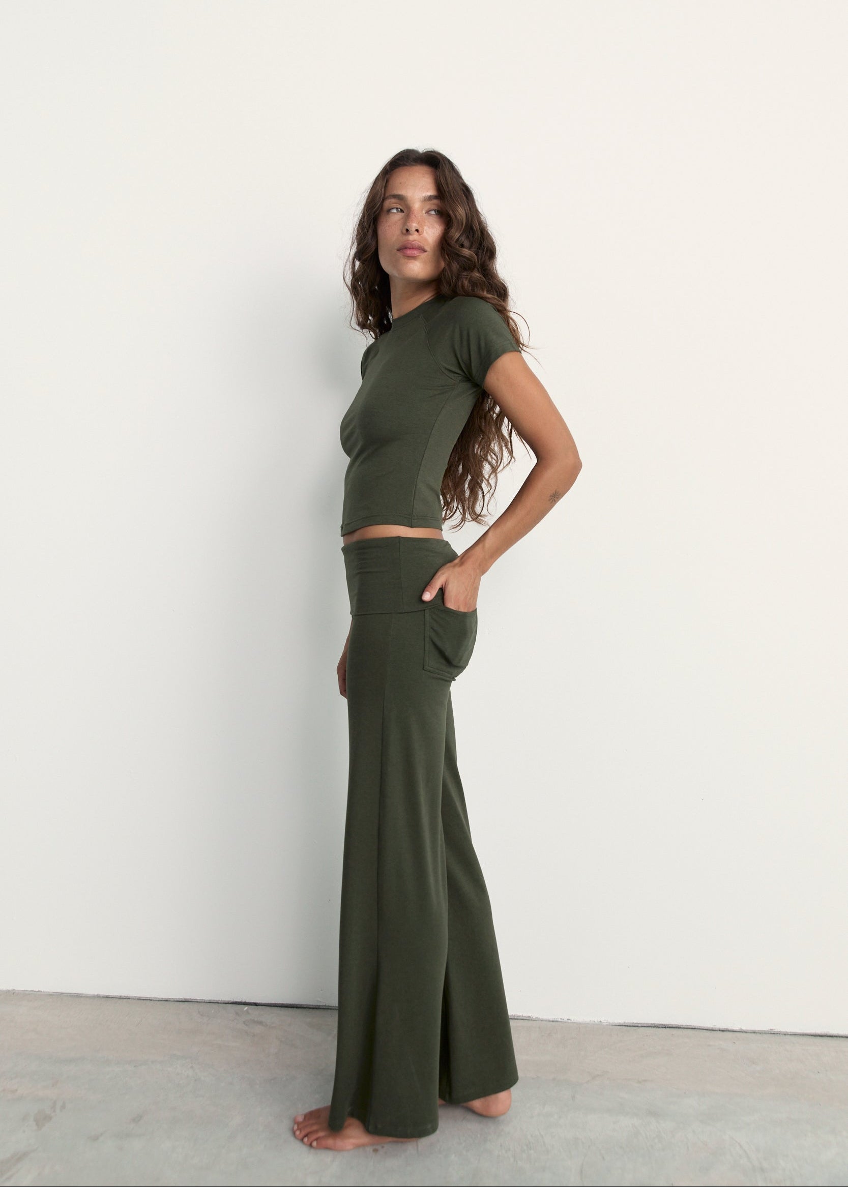A woman modeling an olive green two-piece lounge set consisting of a short-sleeve crop top and matching wide-leg pants, standing against a plain white wall.