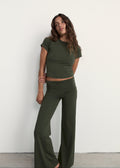 A woman modeling an olive green two-piece lounge set consisting of a short-sleeve crop top and matching wide-leg pants, standing against a plain white wall.