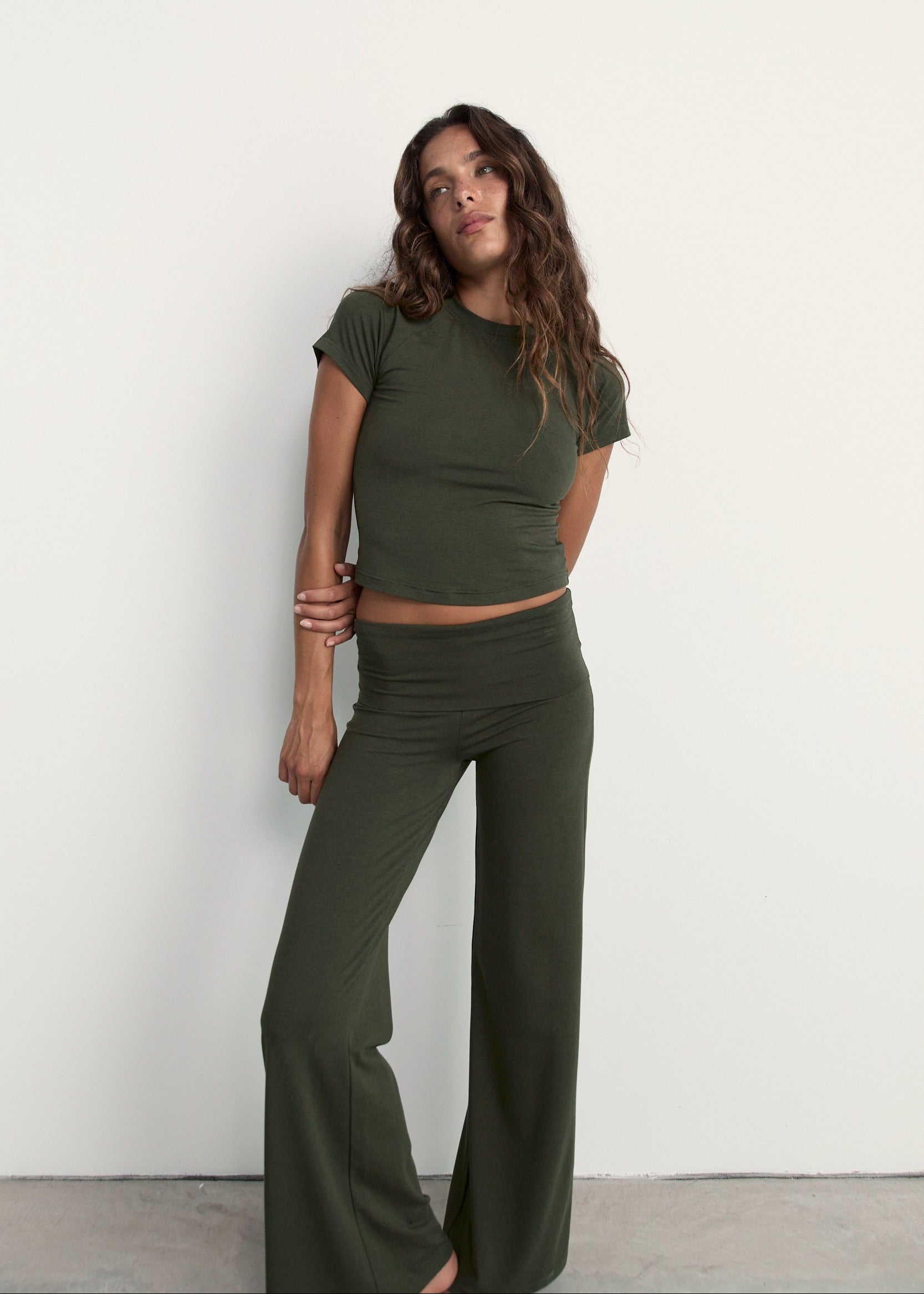 A woman modeling an olive green two-piece lounge set consisting of a short-sleeve crop top and matching wide-leg pants, standing against a plain white wall.