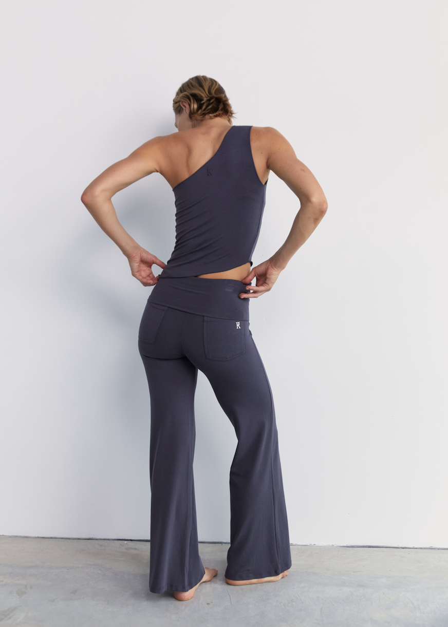 A woman stands with her back to the camera wearing a dark slate gray one-shoulder top and matching high-waisted flare pants against a plain light gray wall.