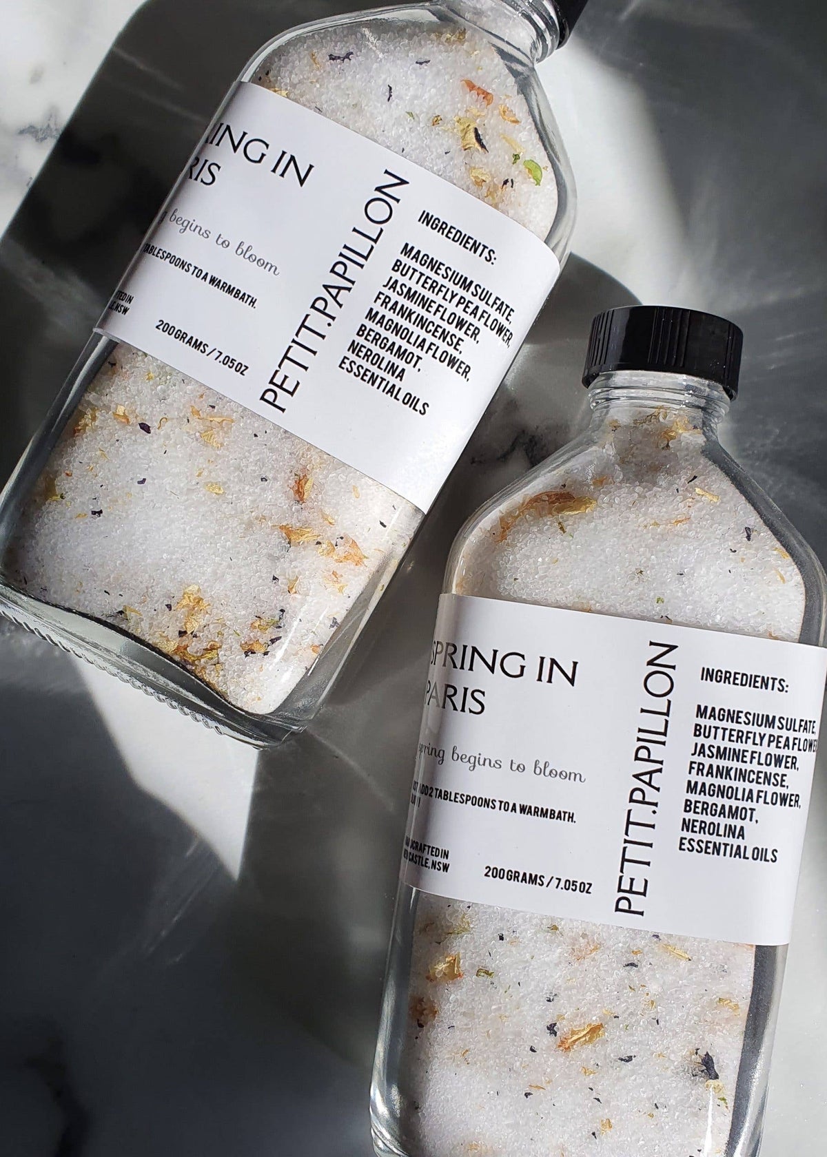 Two clear glass bottles filled with bath salts and dried botanicals, labeled 'PETTIPAPILLON' and 'BRING IN PARIS,' arranged on a marble surface with dramatic shadows.