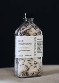 A clear glass bottle with a black cap and a white label reading 'BLUE MOUNTAINS' and 'PETTIPAPILLON', filled with dried botanicals, photographed against a dark background.