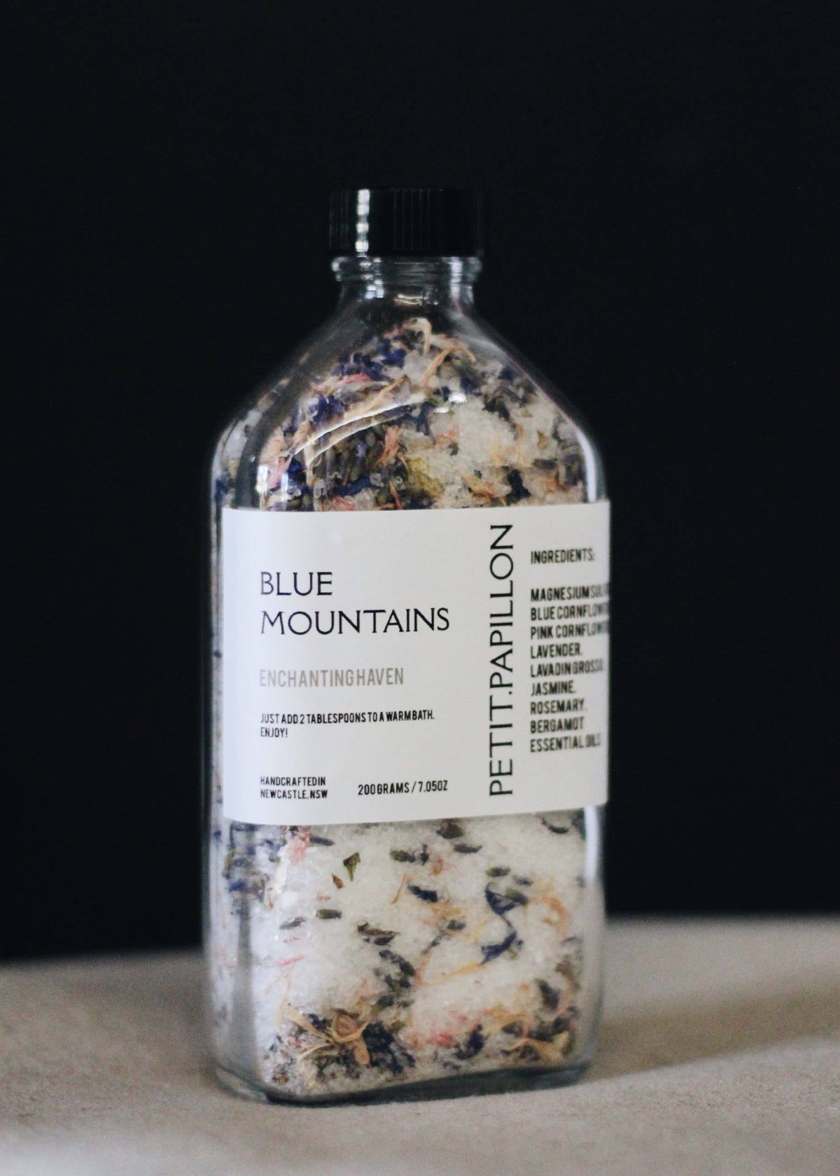 A clear glass bottle with a black cap and a white label reading 'BLUE MOUNTAINS' and 'PETTIPAPILLON', filled with dried botanicals, photographed against a dark background.