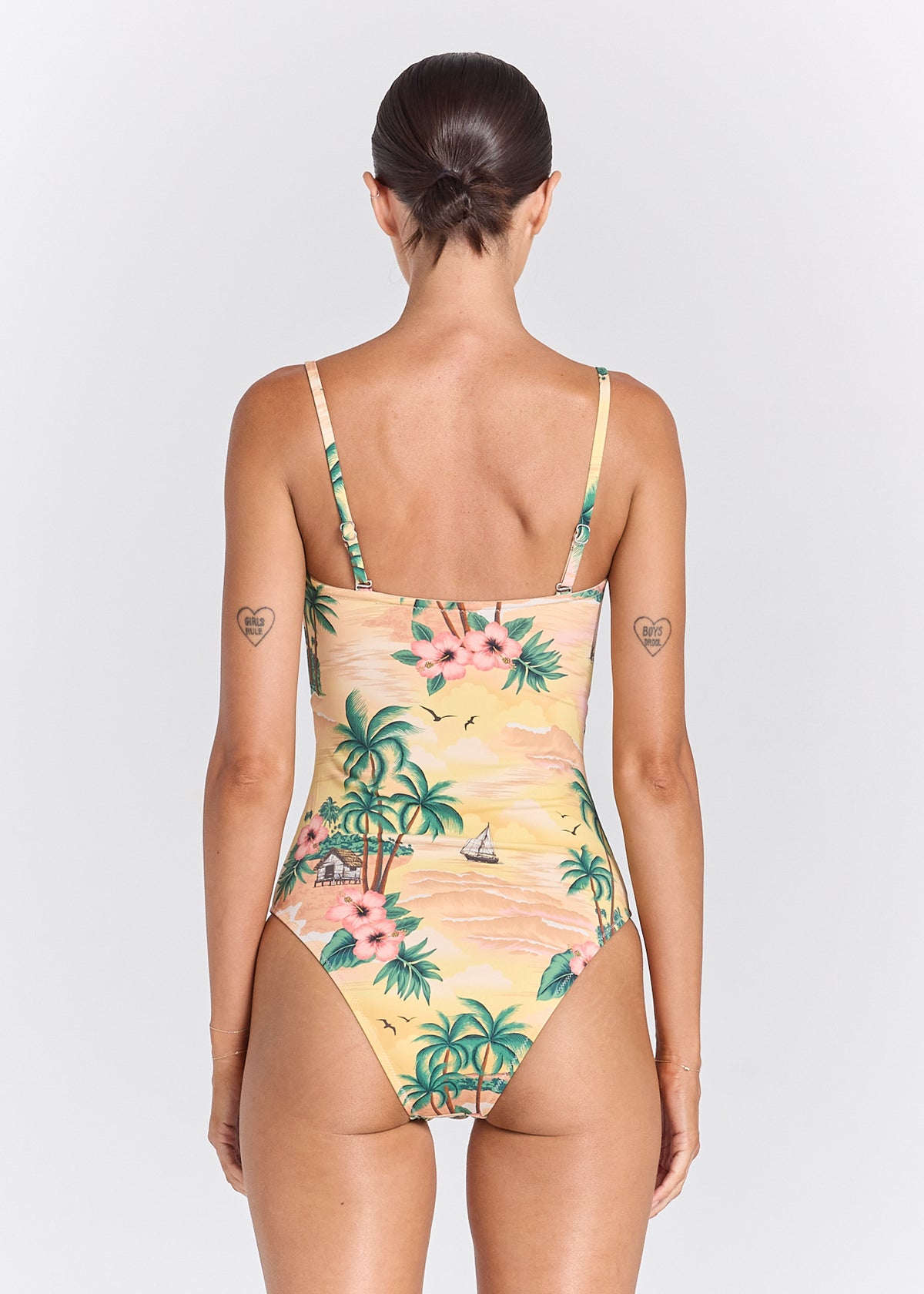 A woman shown from the back wearing a one-piece swimsuit with a tropical beach print on a light yellow base. The print features green palm trees, pink hibiscus flowers, and small sailboats.