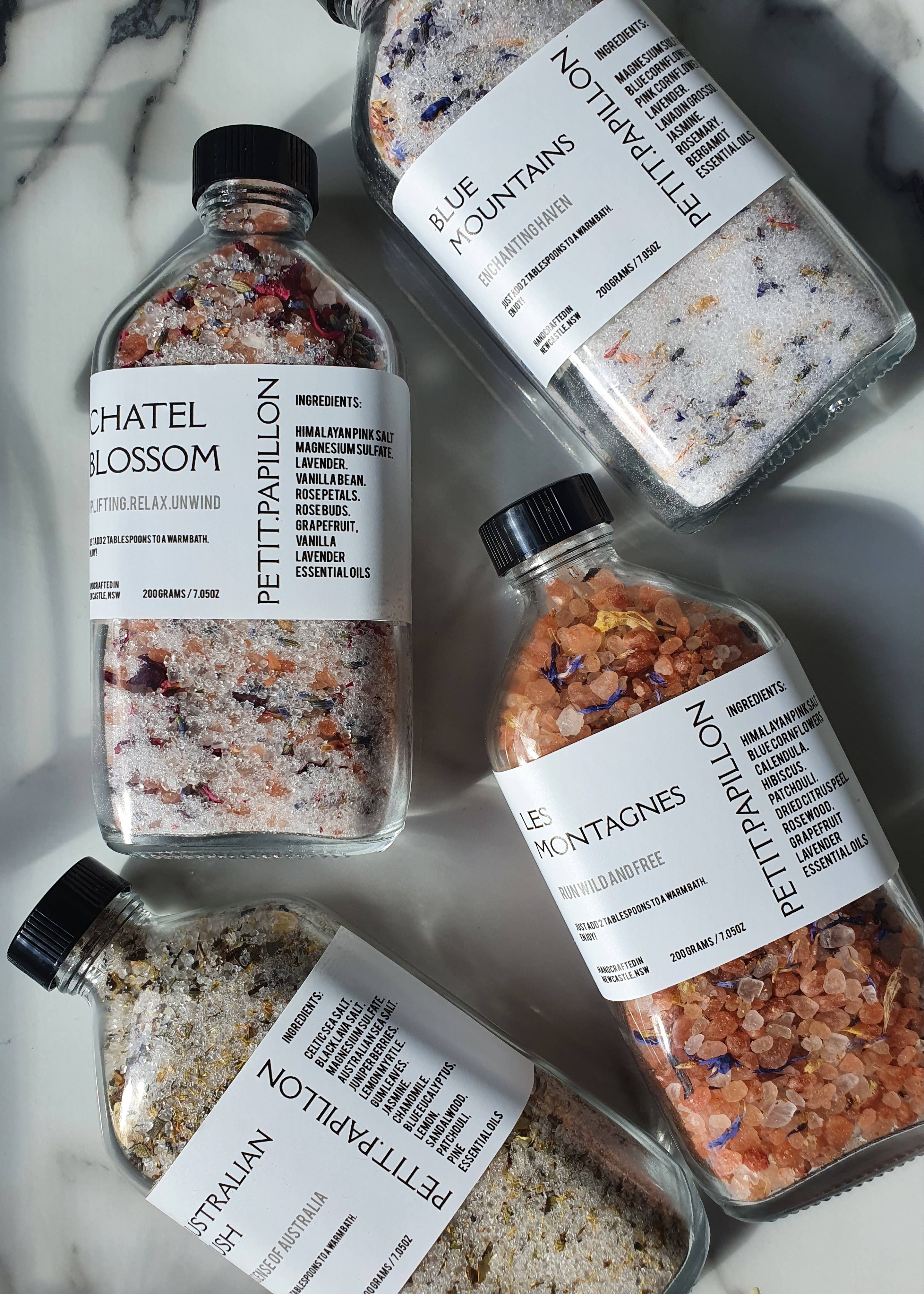 Four clear glass bottles of Pettrapillon bath salts arranged on a white marble surface. The bottles contain different colored salts: pink, blue, green, and orange, with white labels showing the brand name Pettrapillon and product names including Chateau Blossom, Blue Mountains, Australian Gold, and Le Montagnes.
