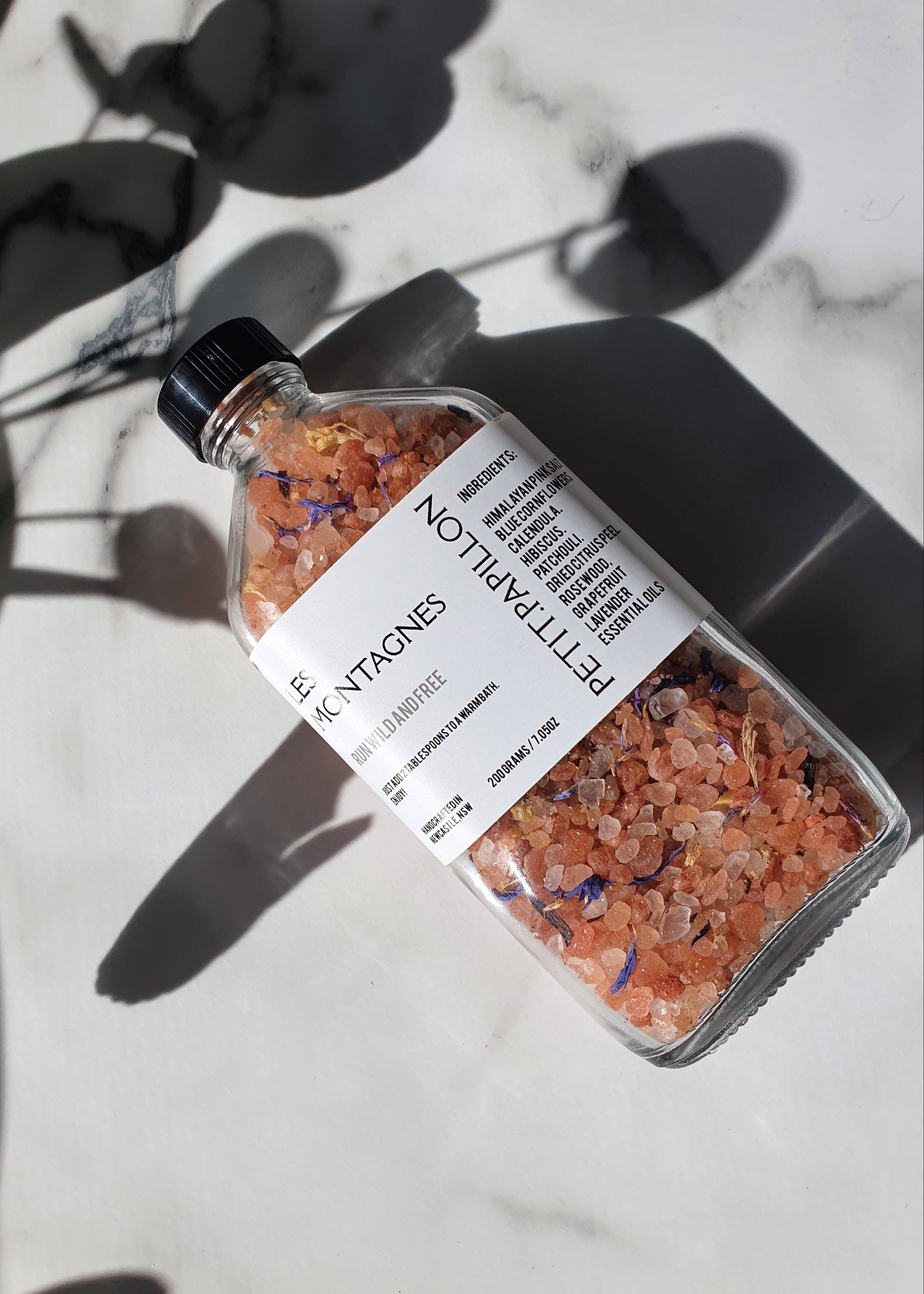 A clear glass bottle filled with pink and orange coarse bath salts, labeled 'PETERPAPILLON' and 'MONTAGNES', placed on a white marble surface with strong leaf shadows.