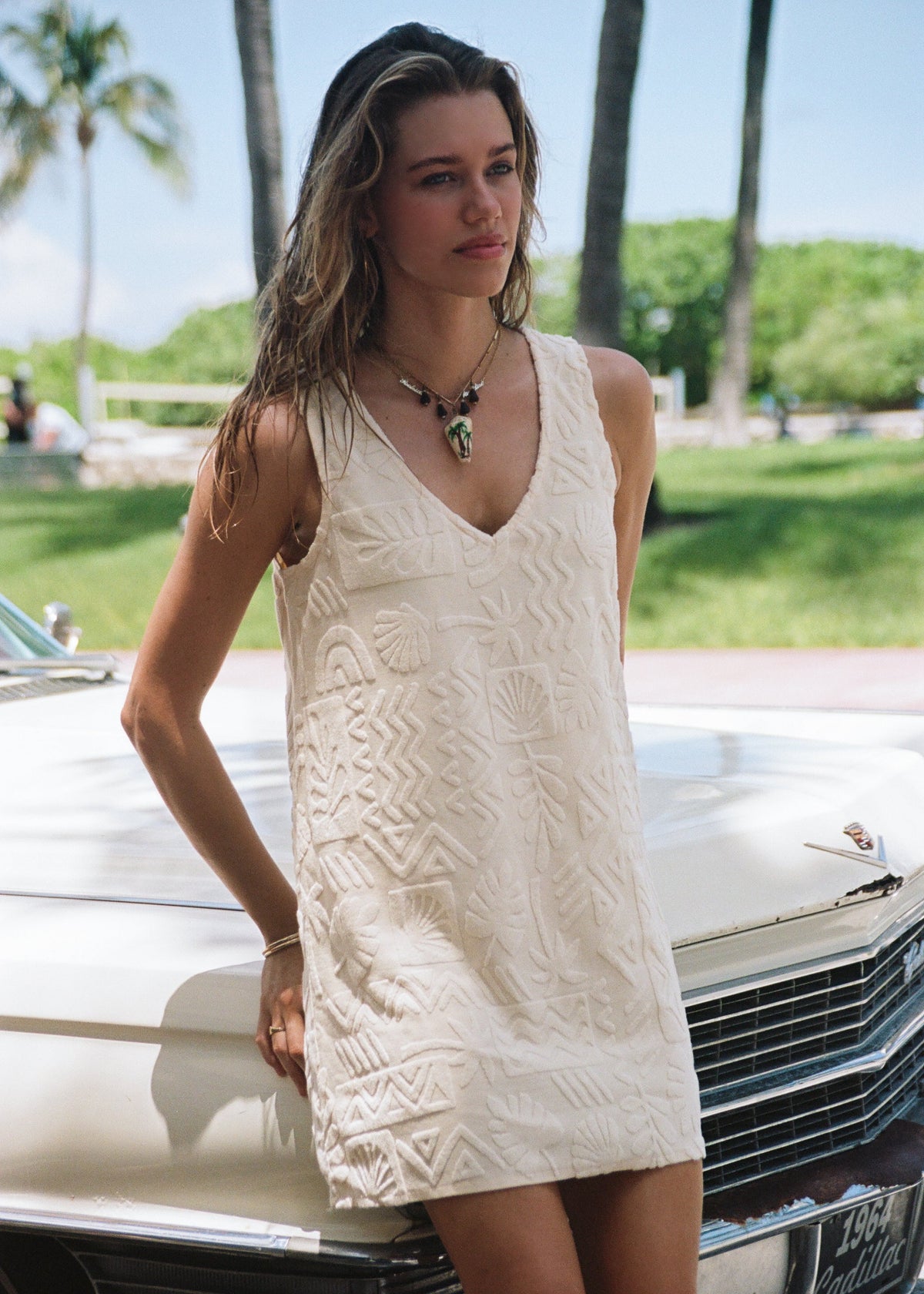 A woman wearing a sleeveless cream-colored textured mini dress leans against a vintage white car outdoors, with palm trees and greenery in the background.
