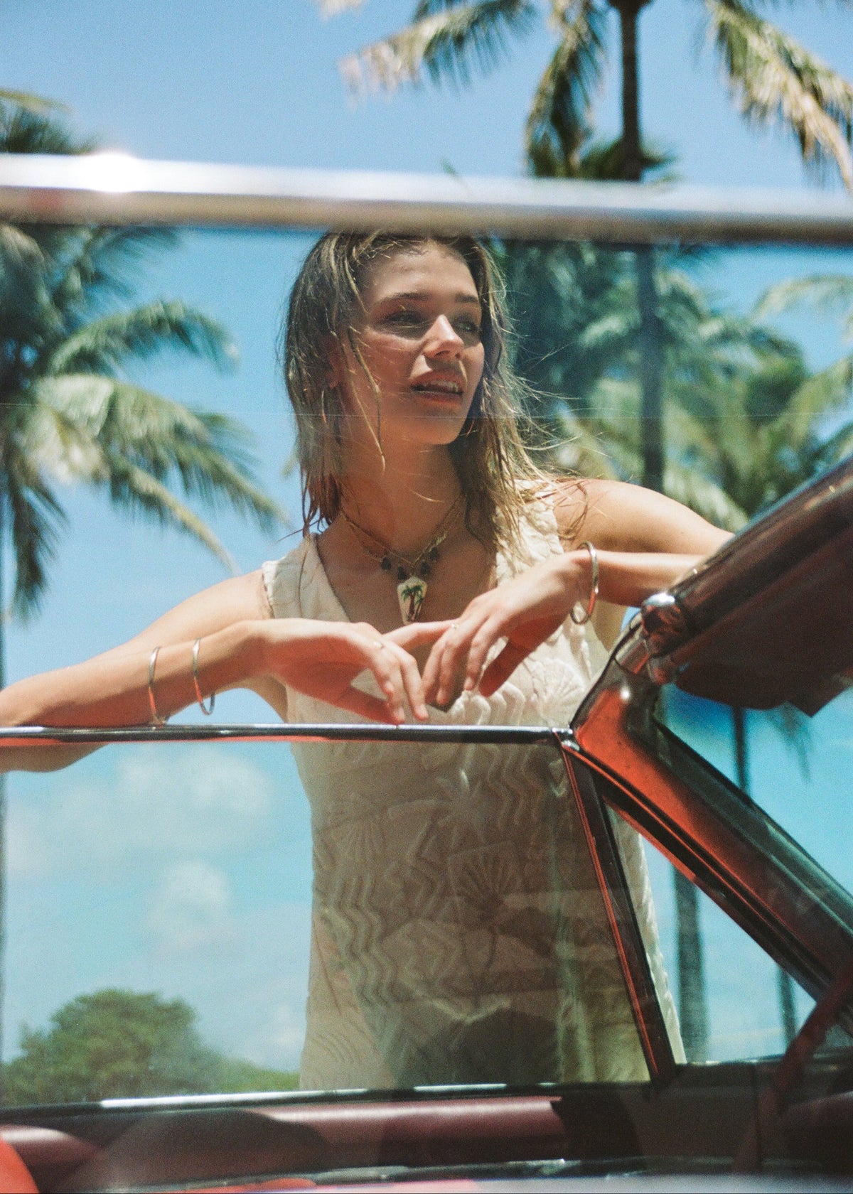 A woman leans over the open window of a vintage car outdoors, with palm trees and blue sky in the background. She is wearing a light beige sleeveless top and layered necklaces.
