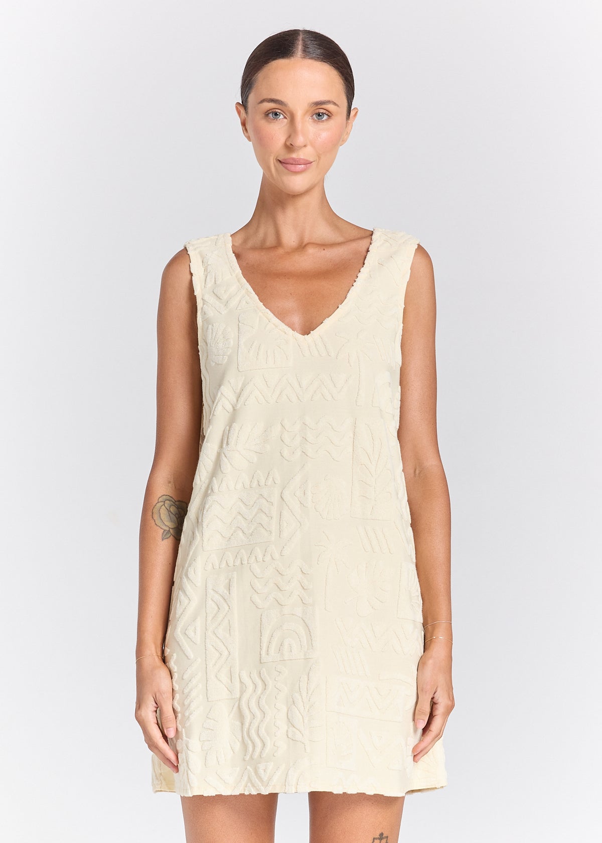 A woman modeling a sleeveless, V-neck cream-colored dress with a textured, geometric patterned design, set against a plain light gray studio background.