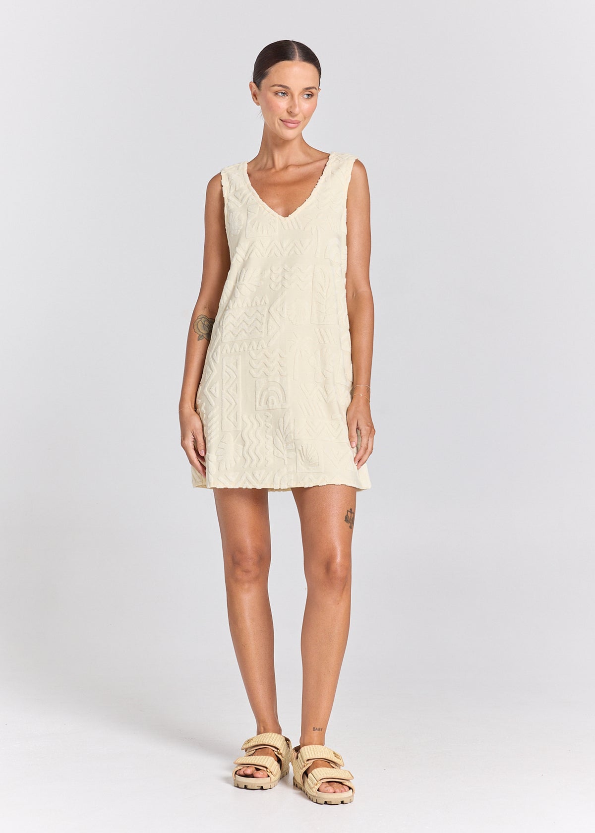 A woman modeling a sleeveless, V-neck cream-colored knit dress with a textured cable pattern, paired with beige double-strap sandals, against a light gray studio background.