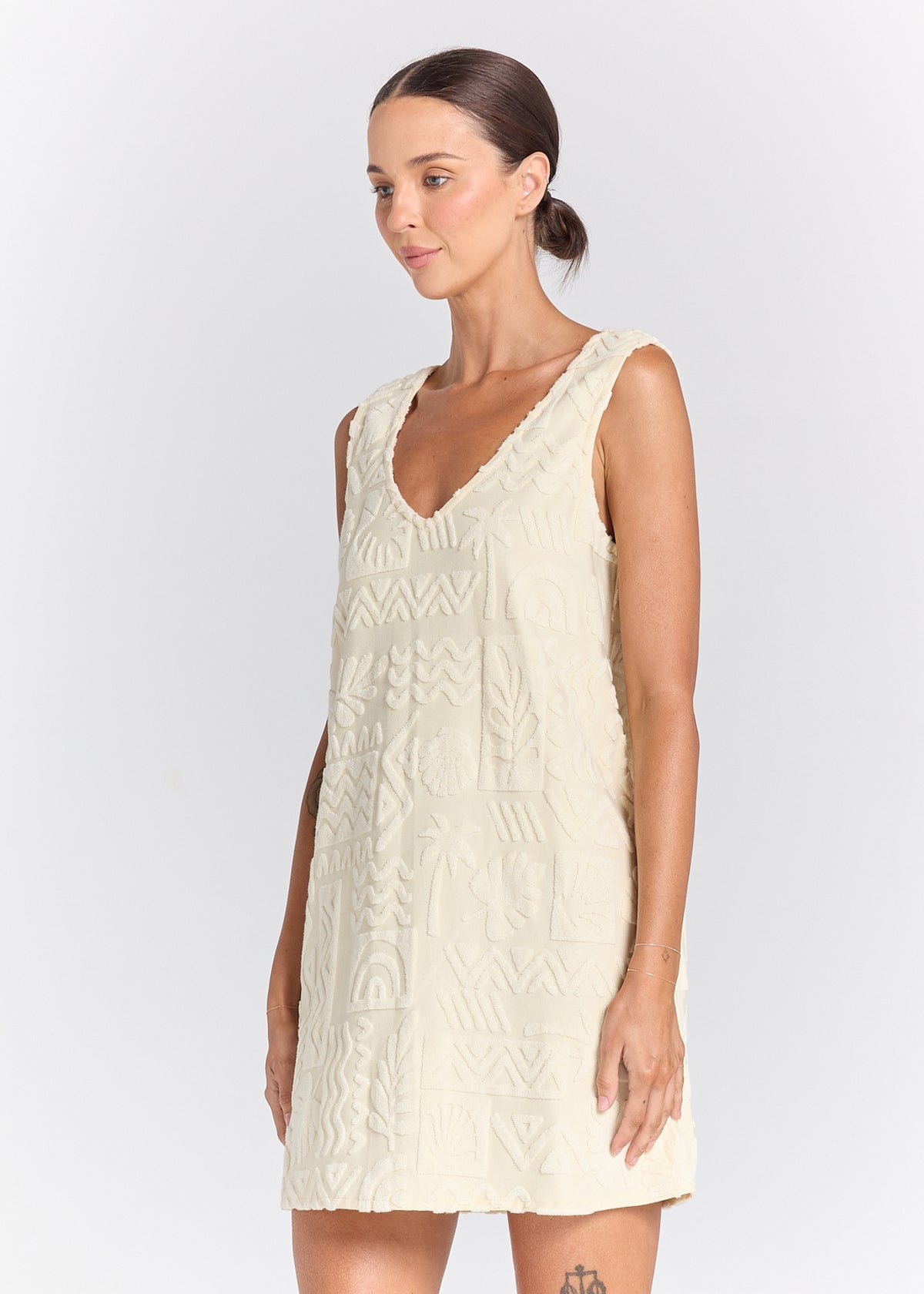 A woman modeling a sleeveless cream-colored dress with a textured, geometric embroidered pattern and a scalloped V-neckline, set against a plain light gray studio background.