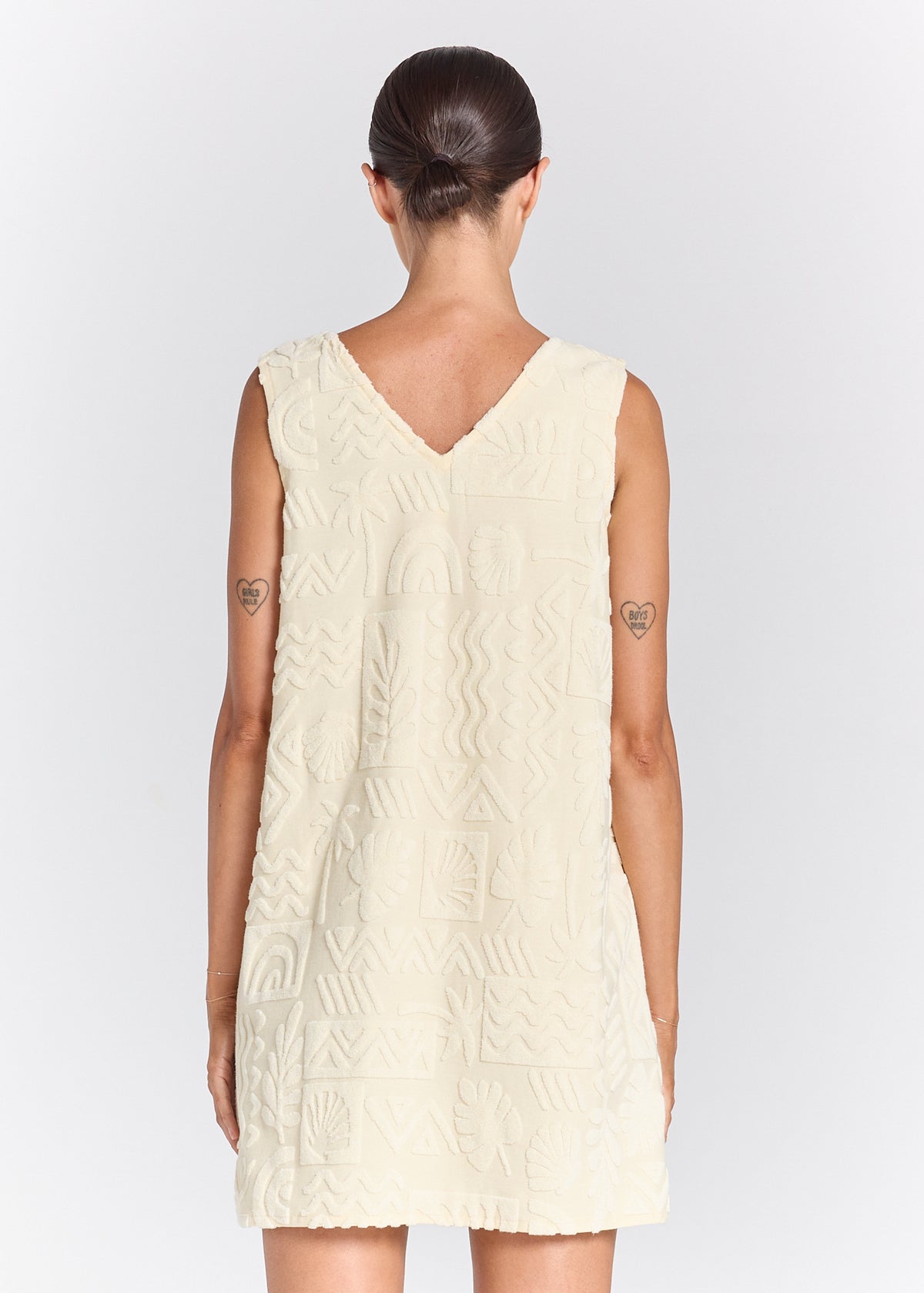A woman shown from the back wearing a sleeveless cream-colored dress with a textured geometric pattern and a V-neckline, set against a plain light gray background.