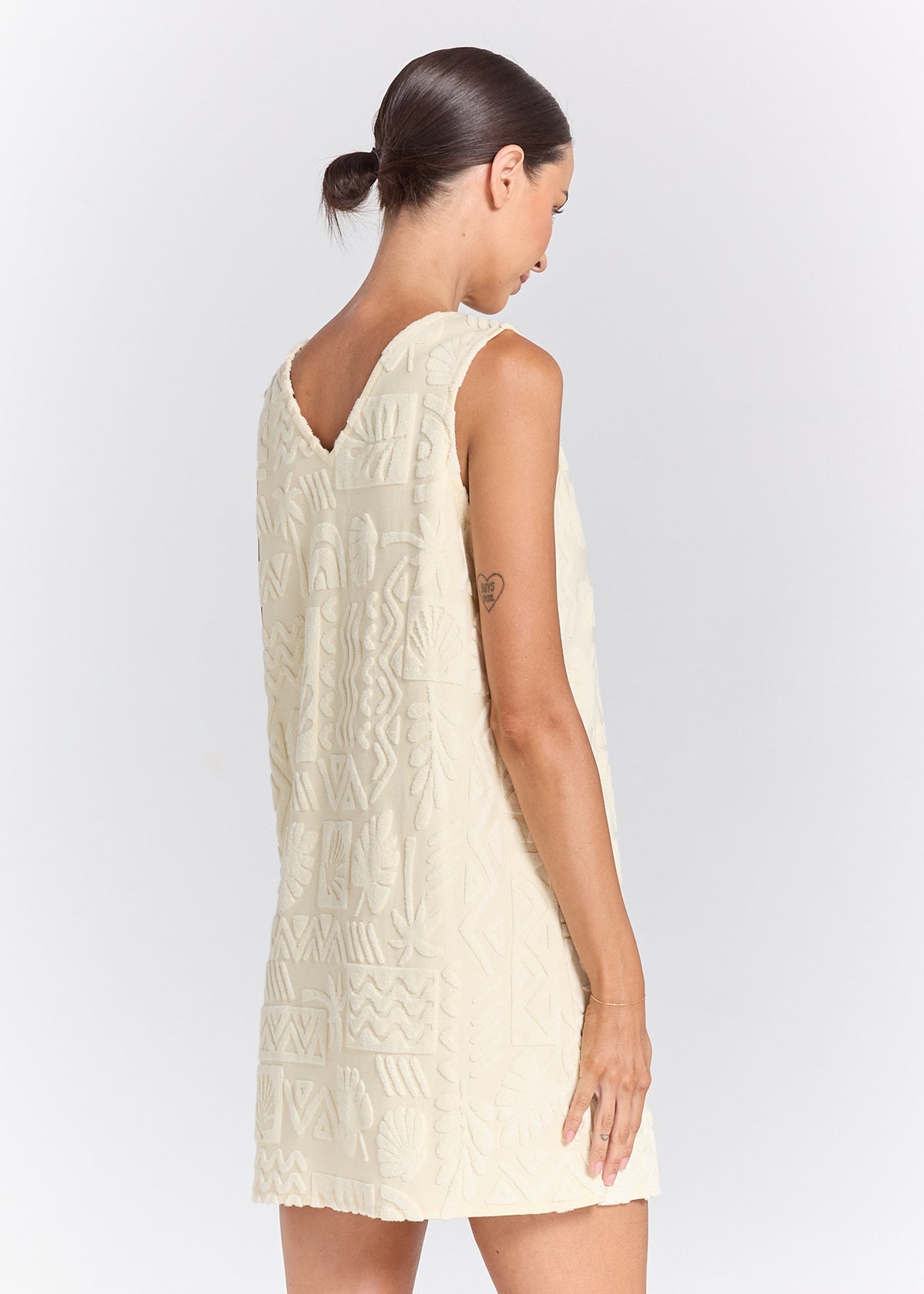 A woman shown from the back wearing a sleeveless cream-colored dress with a textured geometric pattern, photographed against a light gray studio background.