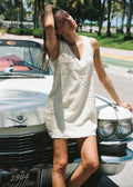 A woman wearing a white textured sleeveless mini dress leans against a vintage white Cadillac convertible outdoors on a sunny street with palm trees in the background.