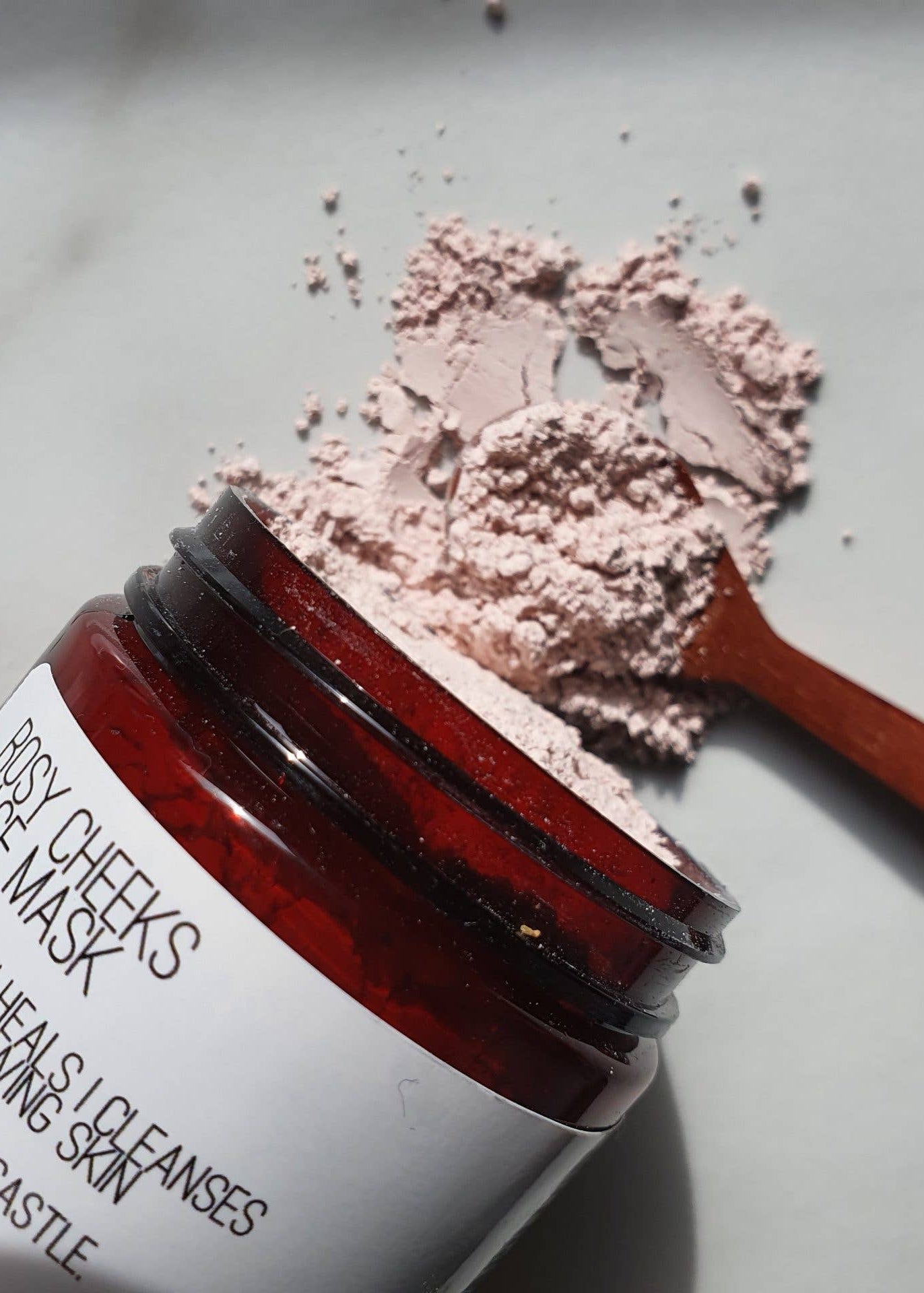 An open amber glass jar of Rose Cheeks Face Mask with a white label, shown on a light marble surface with the pink cleanser powder spilled above and a small wooden spoon holding some of the mask.