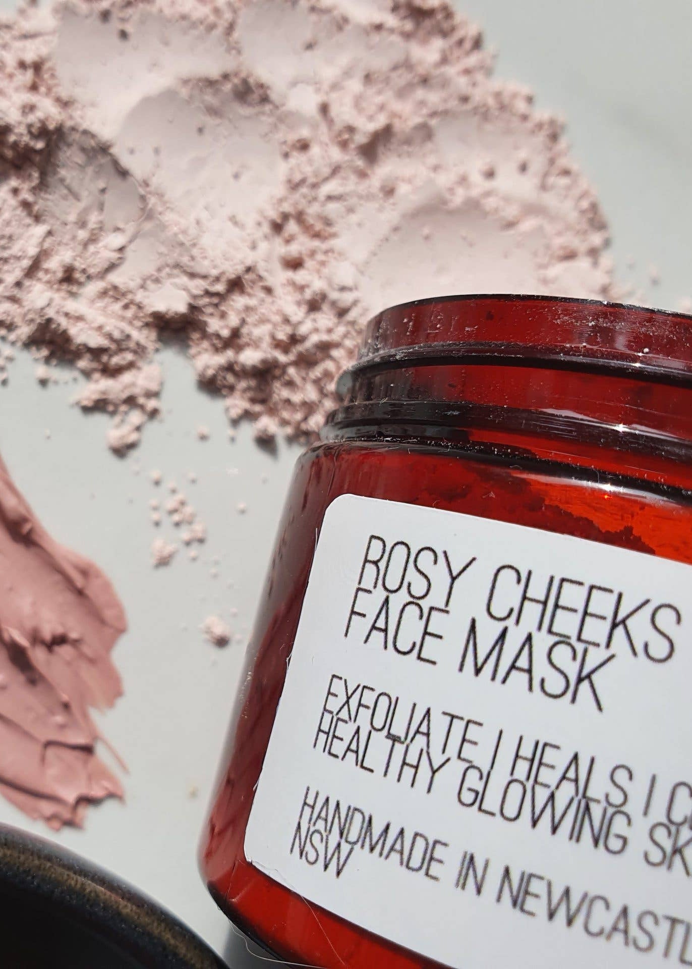 An amber glass jar labeled 'ROSY CHEEKES FACE MASK' with a white paper label, shown next to a thick pinkish-rose face mask smear and loose mask powder on a light marble surface.