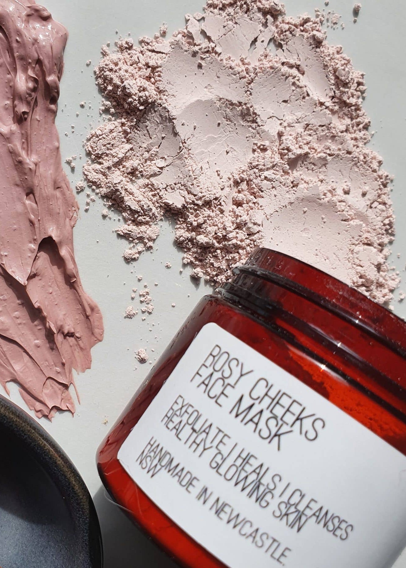 A jar of Rosy Cheeks Face Mask powder shown on a light marble surface with the pink powder smeared and sprinkled beside it, plus a small wooden spoon in a dark bowl.