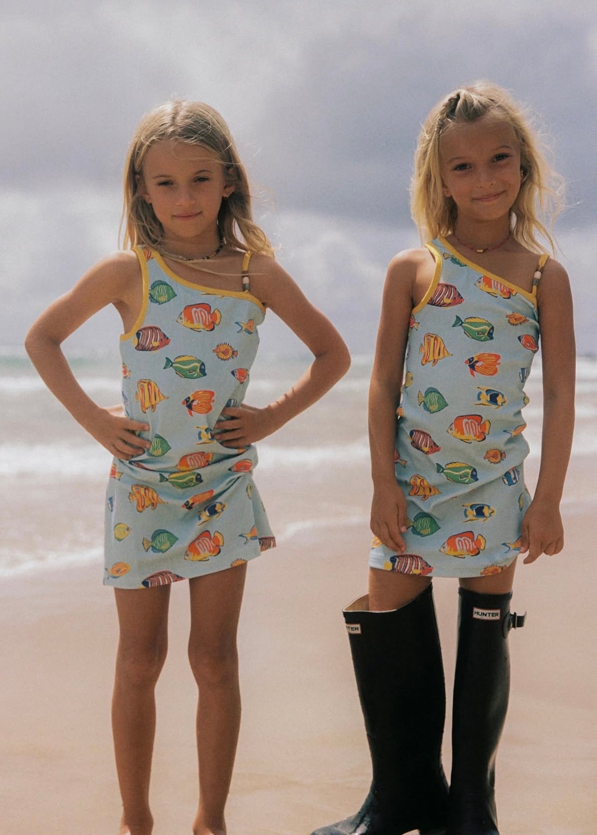 Two young girls standing on a sandy beach wearing matching one-shoulder mini dresses with a tropical fish print on a light blue background. The girl on the left wears no boots, while the girl on the right wears tall black rain boots.