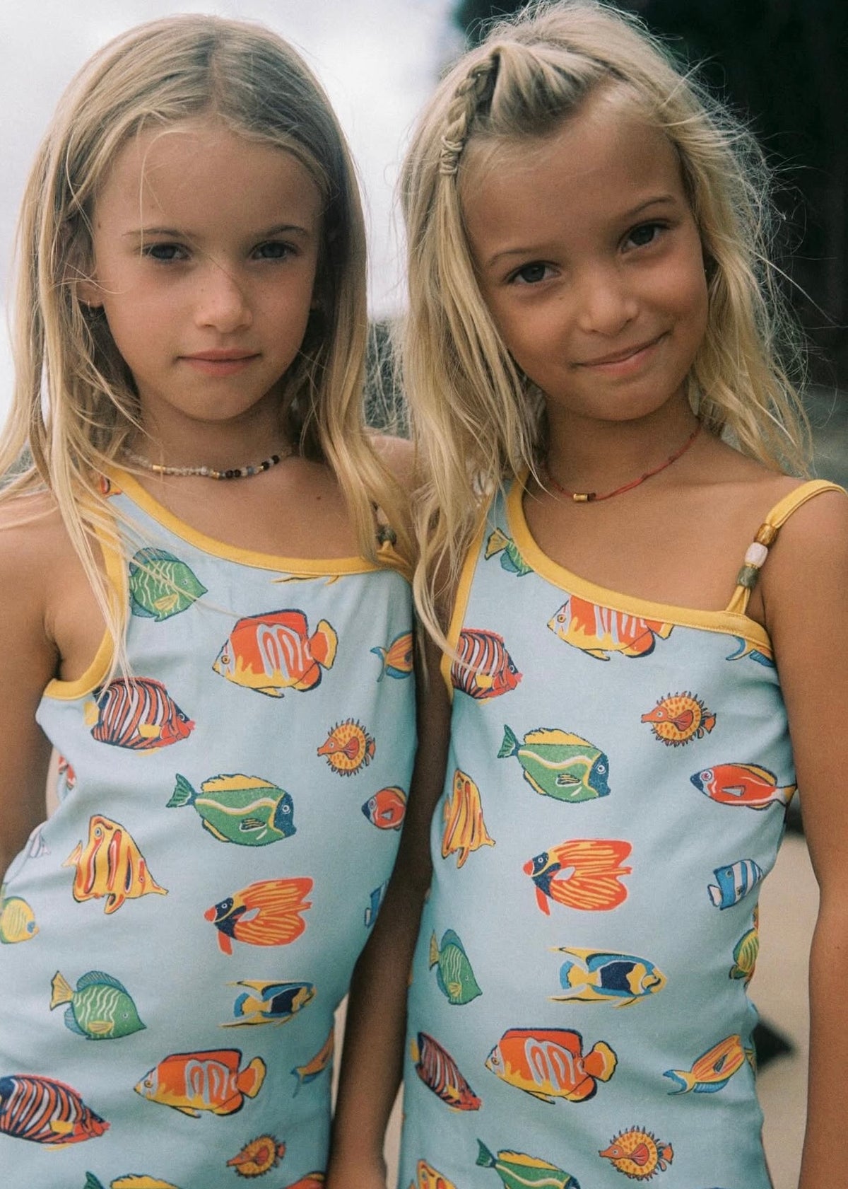 Two young girls wearing matching light blue tank tops with a colorful tropical fish print and yellow trim, standing outdoors.