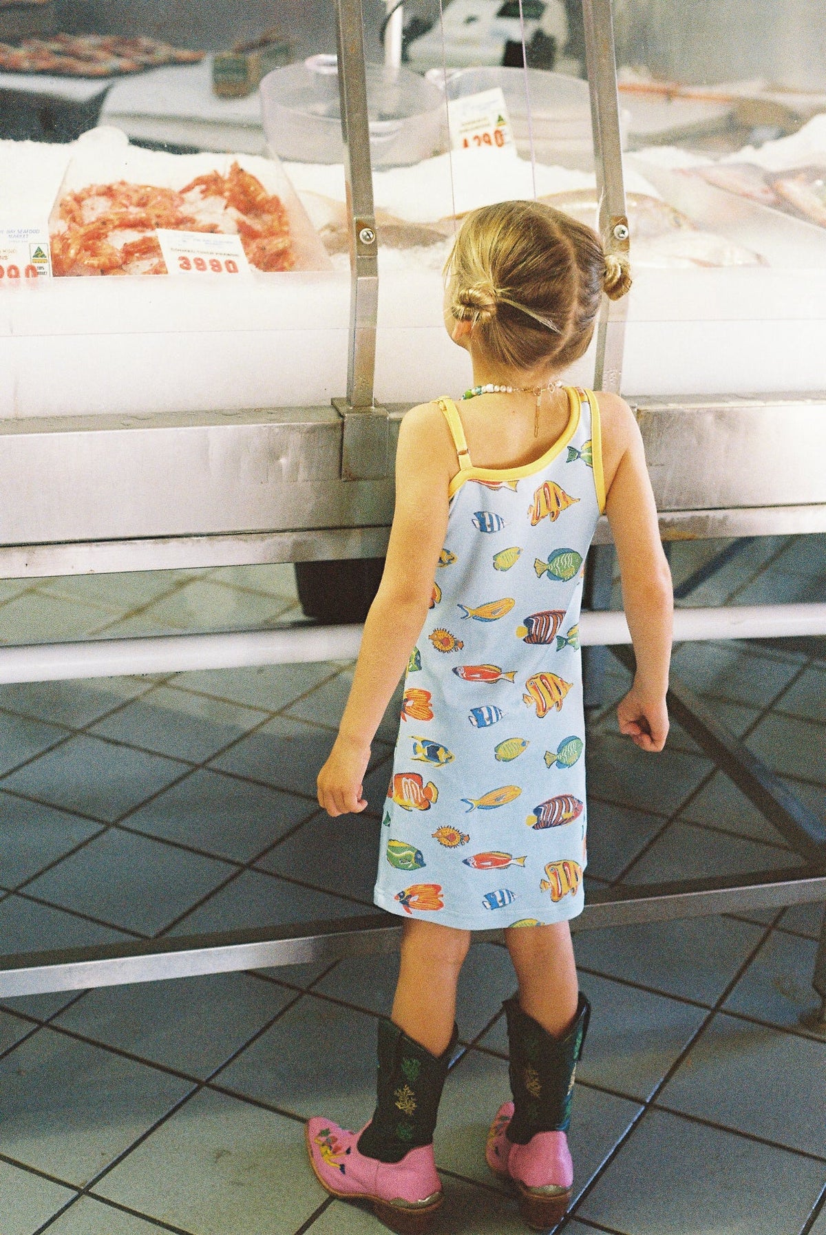 Child in a patterned dress and cowboy boots standing in front of a counter with food items.