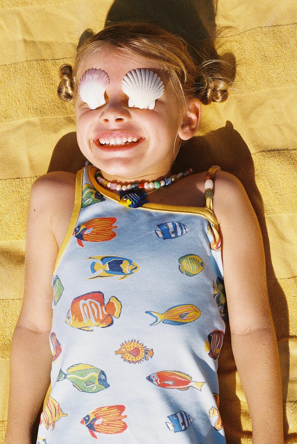 Child wearing a colorful fish-patterned swimsuit with shell sunglasses on a yellow towel.
