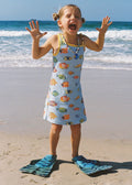 A young girl stands on a sandy beach with the ocean in the background, wearing a light blue sleeveless dress covered in colorful fish prints and blue flip-flops.