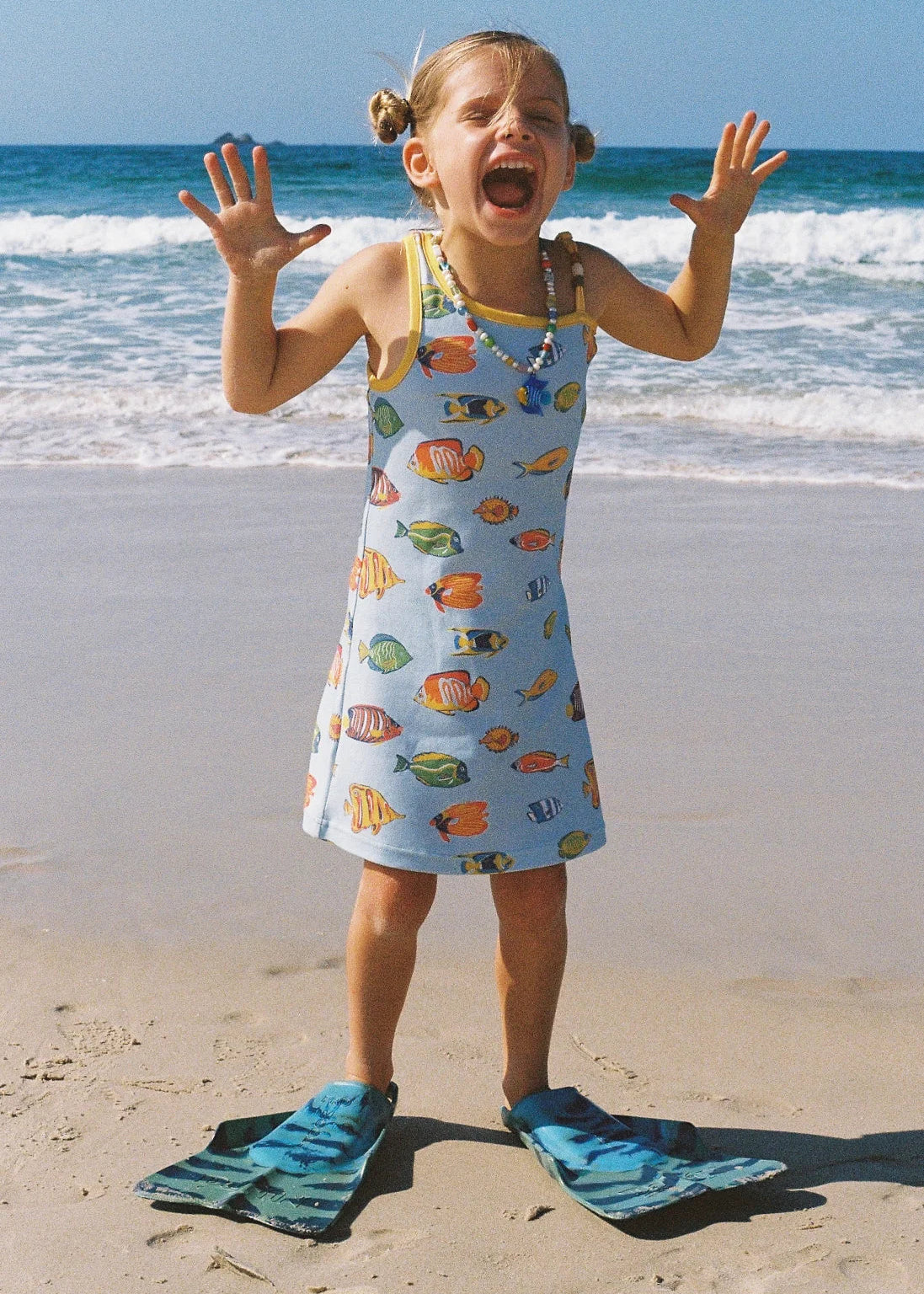 A young girl stands on a sandy beach with the ocean in the background, wearing a light blue sleeveless dress covered in colorful fish prints and blue flip-flops.