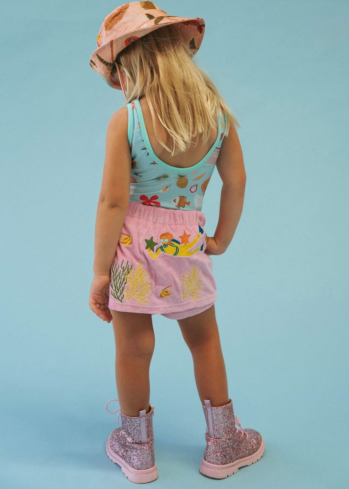 A young girl is shown from behind wearing a matching two-piece swimsuit set with a light blue top and pink shorts, both decorated with colorful sea-themed illustrations. She is also wearing a pink patterned sun hat and pink glittery ankle boots, standing against a solid light blue background.