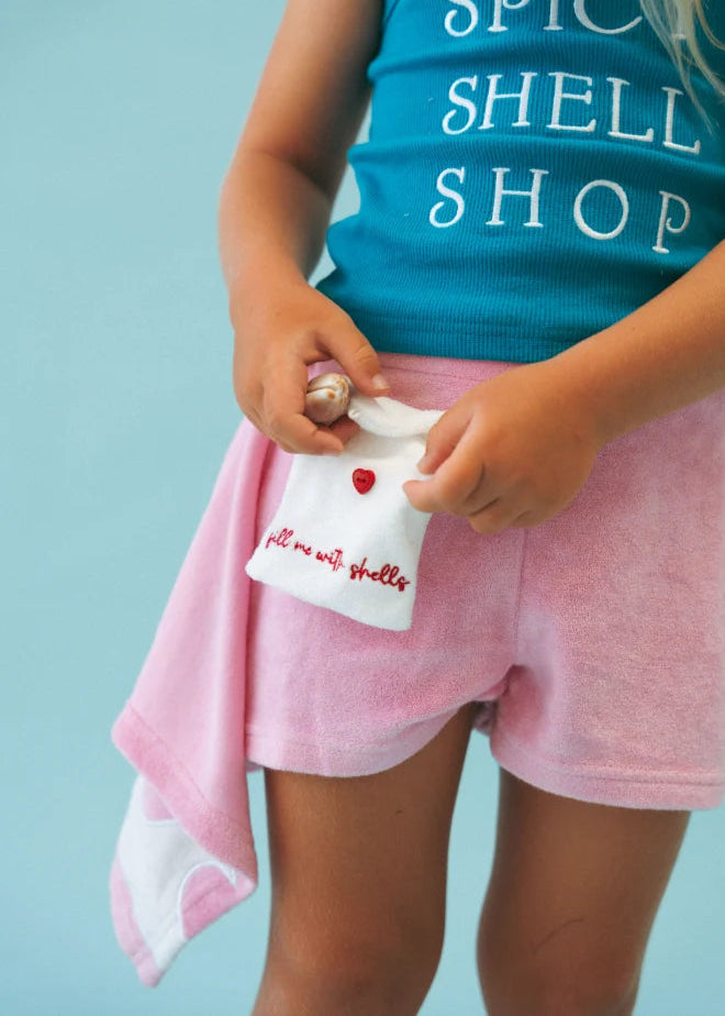 A child wearing a turquoise tank top that says 'SPICY SHELL SHOP' and pink shorts, holding a small white pouch with red script and a red heart on a light blue background.