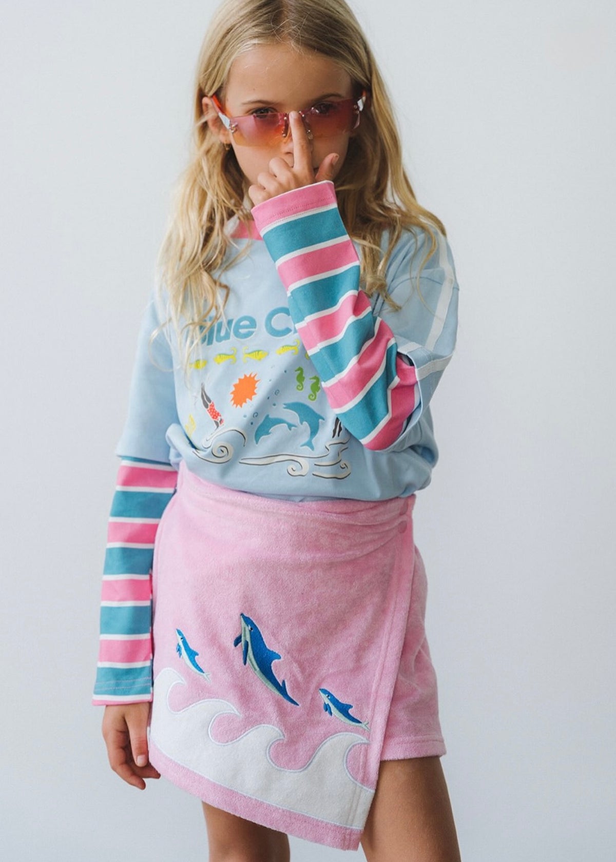 A young girl wearing a light blue long-sleeve graphic sweatshirt with pink and teal striped sleeves, paired with a pink and white towel skirt featuring blue dolphin prints, set against a plain light gray background.