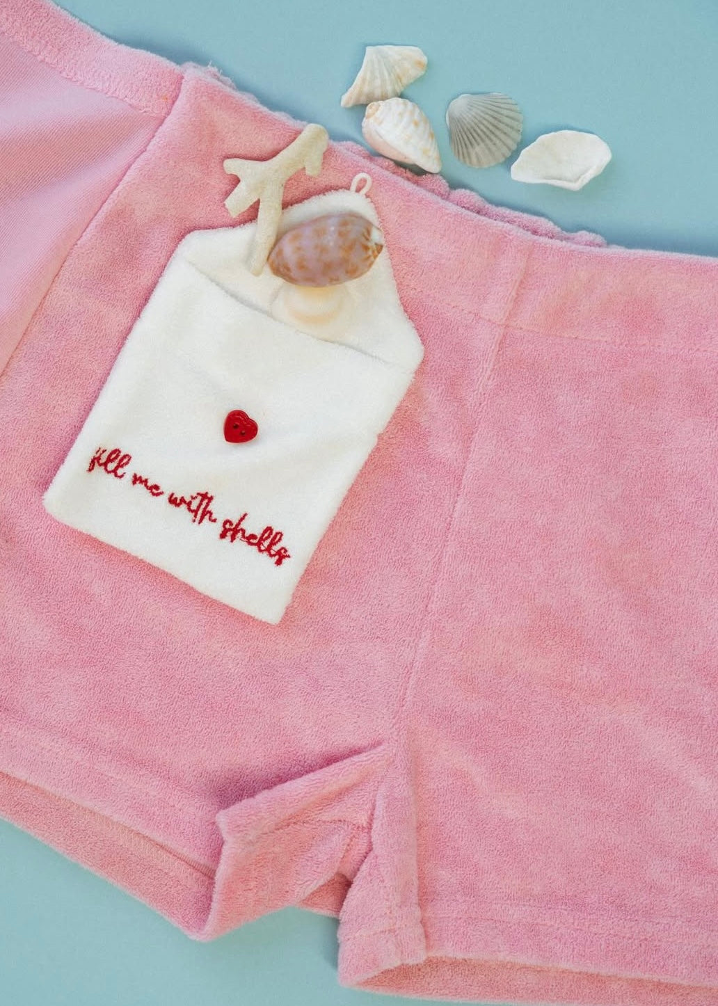 Pink plush shorts with a white embroidered pocket that reads 'fill me with shells' and a small red heart, styled with seashells on a light blue background.