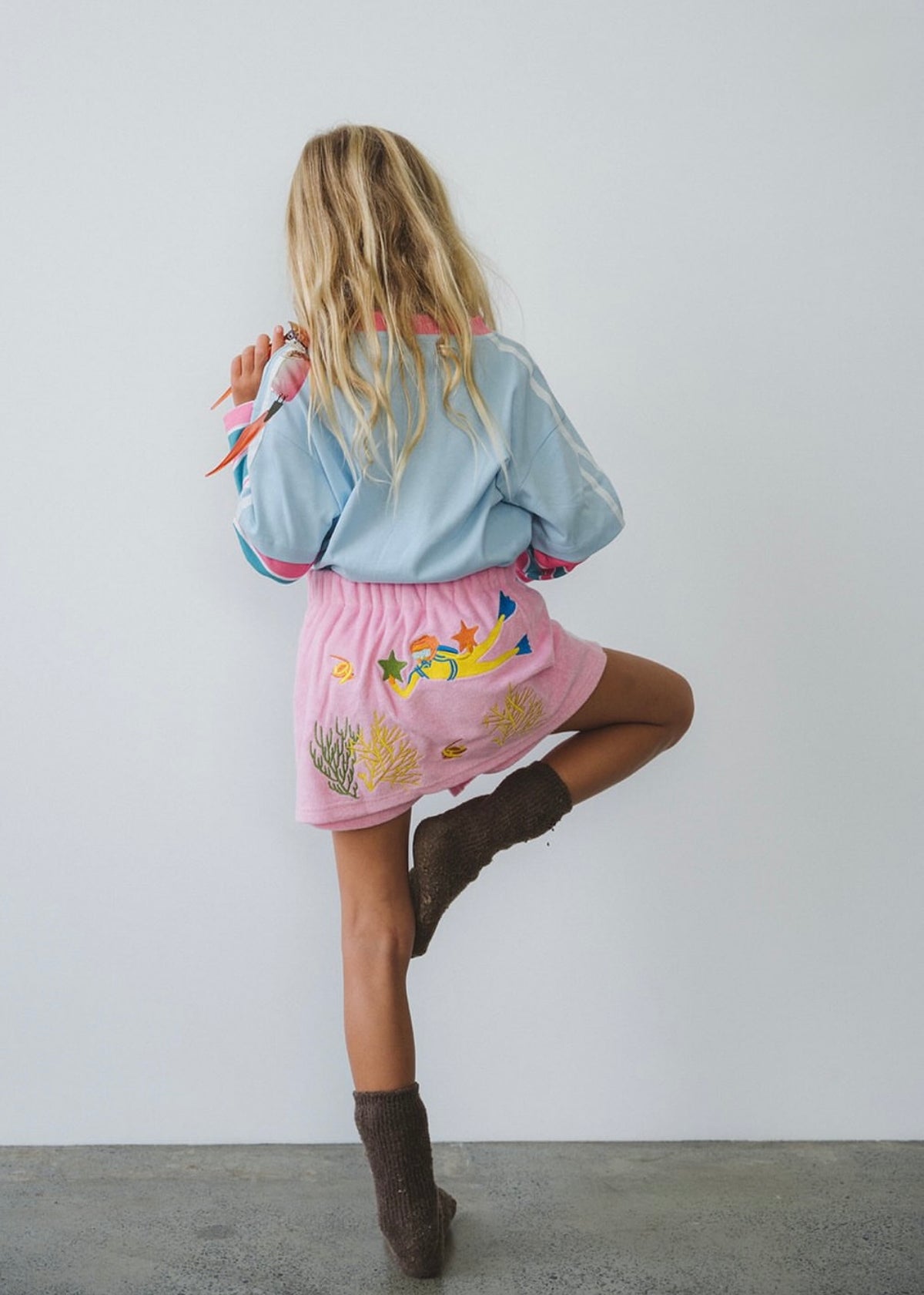 A young girl is shown from behind wearing a light blue long-sleeve shirt and a pink skirt with colorful sea-creature embroidery, paired with brown boots, posing against a plain white wall.