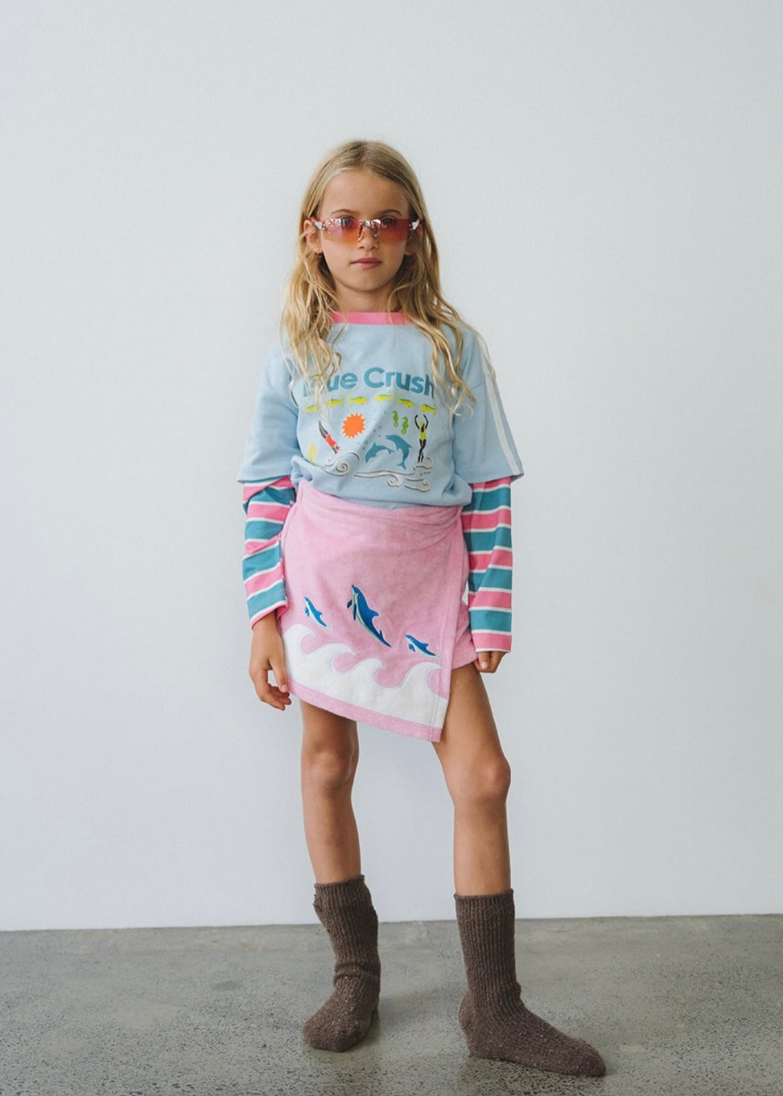 A young girl stands in a studio wearing a light blue graphic long-sleeve shirt, a pink striped and cloud-print mini skirt, and brown socks. She is wearing pink-tinted sunglasses against a plain white wall and concrete floor.