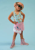 A young girl modeling a light blue tank top with a tropical print, a pink towel-style skirt with a shark design, a pink patterned hat, black sunglasses, and pink lace-up boots, posed against a solid light blue background.