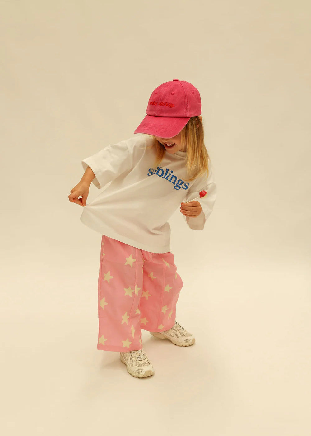 Child wearing a white shirt with text, pink pants, and a red cap on a beige background