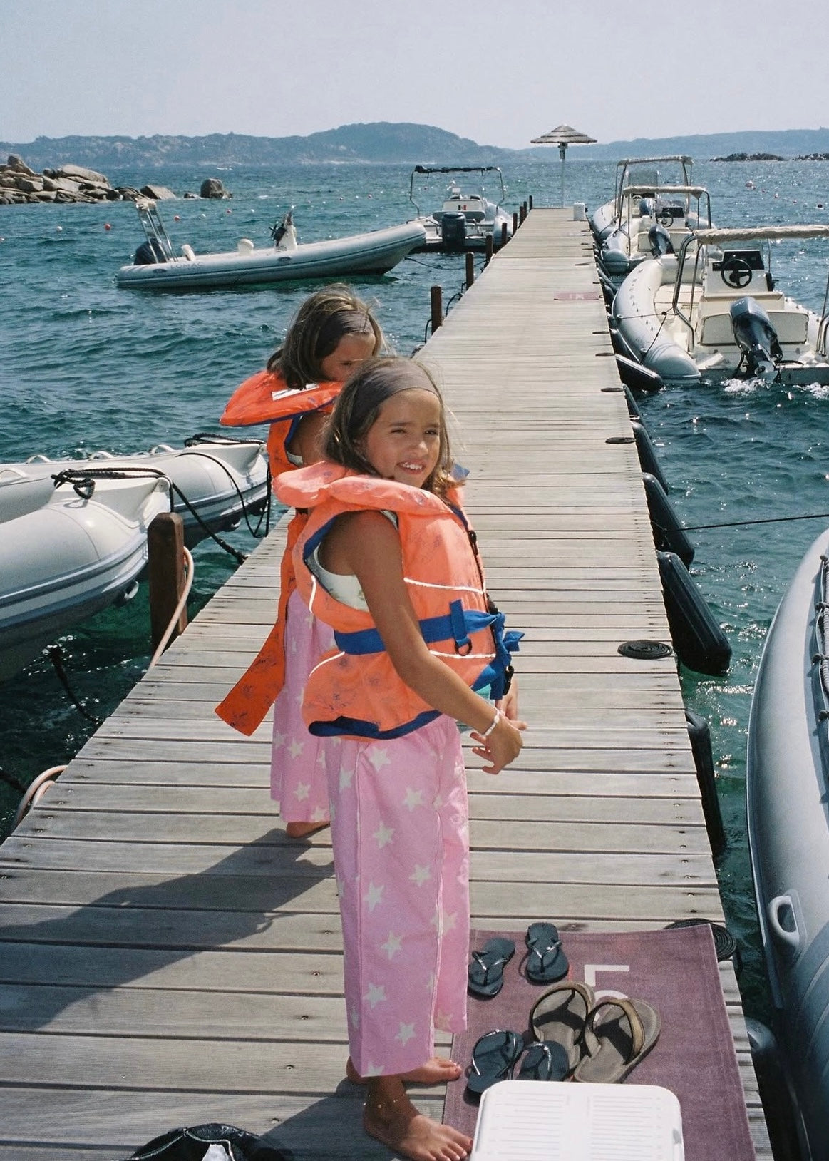 Two young girls standing on a wooden dock at a marina, wearing orange life jackets. Inflatable boats are docked on both sides of the wooden pier, with water and distant hills in the background.