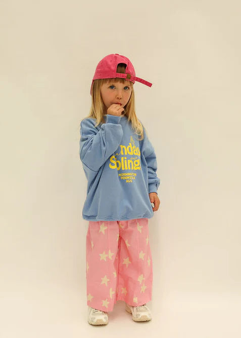 Child wearing a blue sweatshirt and pink pants with a red cap on a beige background