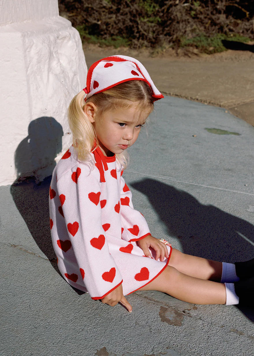 Young girl sitting on a stone surface wearing a red and white dress with heart patterns.