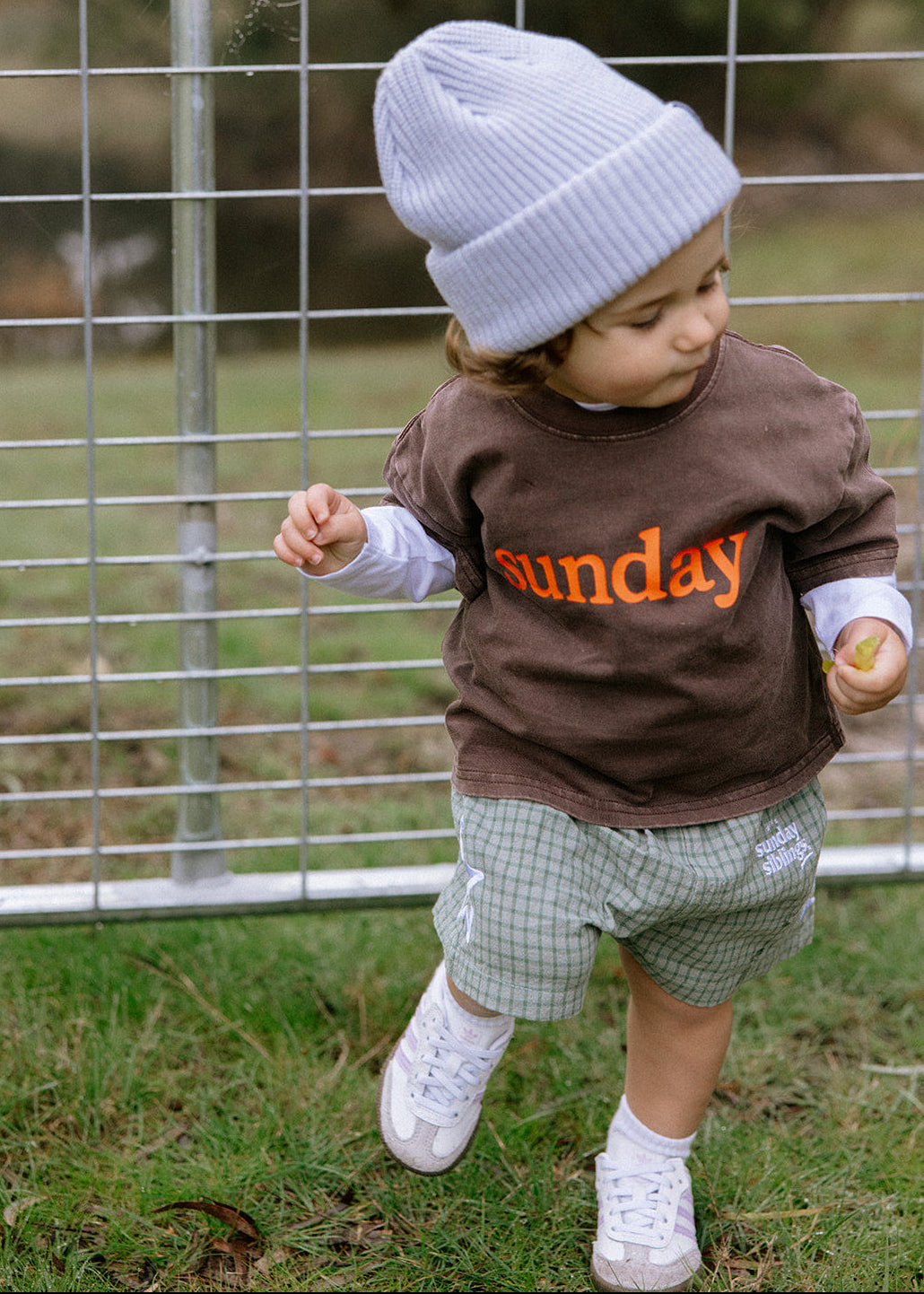 A toddler wearing a brown crewneck sweatshirt with 'sunday' printed in orange, a light blue knit beanie, and green plaid shorts, walking outdoors in front of a wire fence.