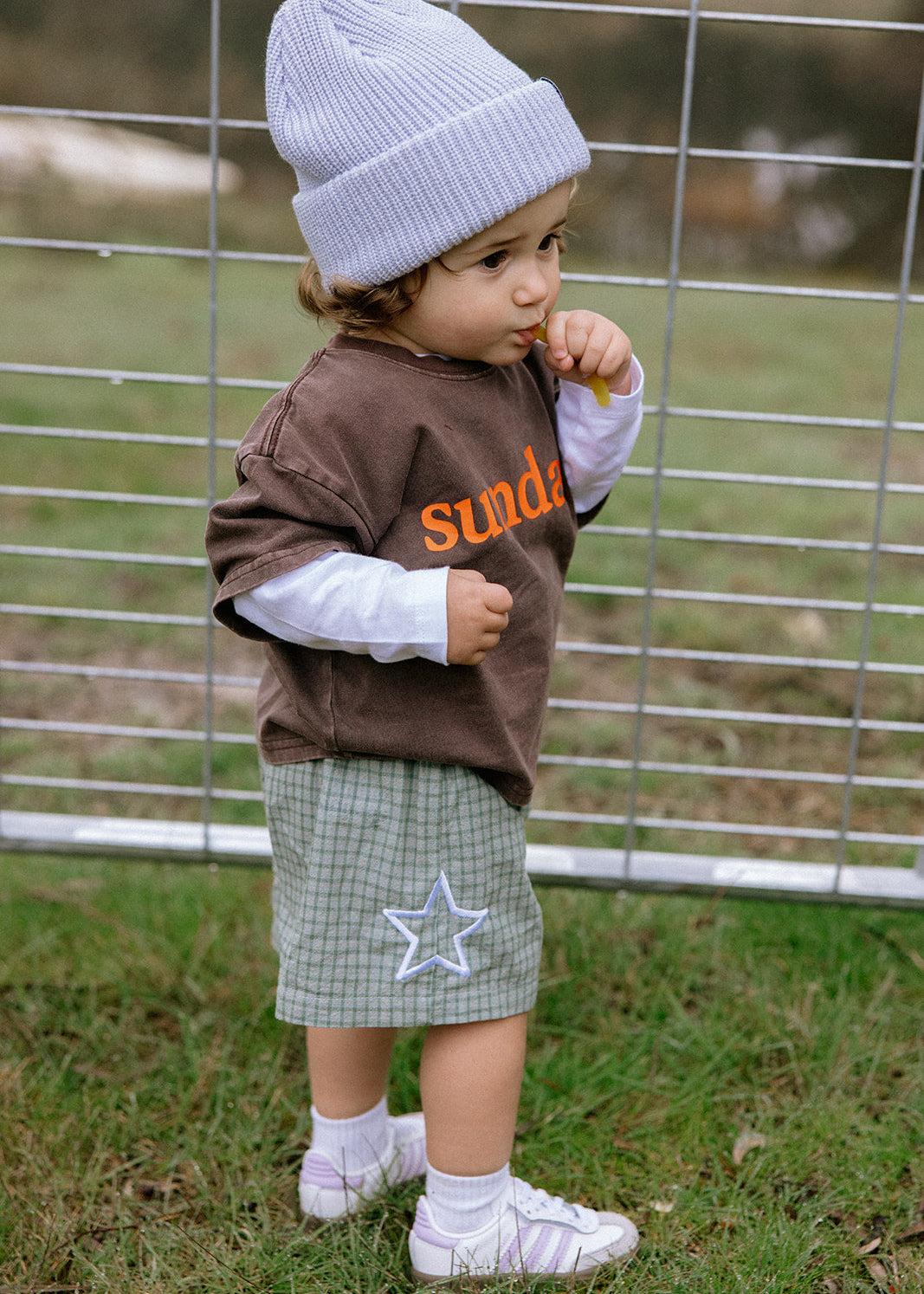 A toddler standing outdoors in front of a wire fence, wearing a light blue knit beanie, a brown long-sleeve shirt with orange text over a white long-sleeve layer, a green plaid skirt with a white star applique, white socks, and white shoes.