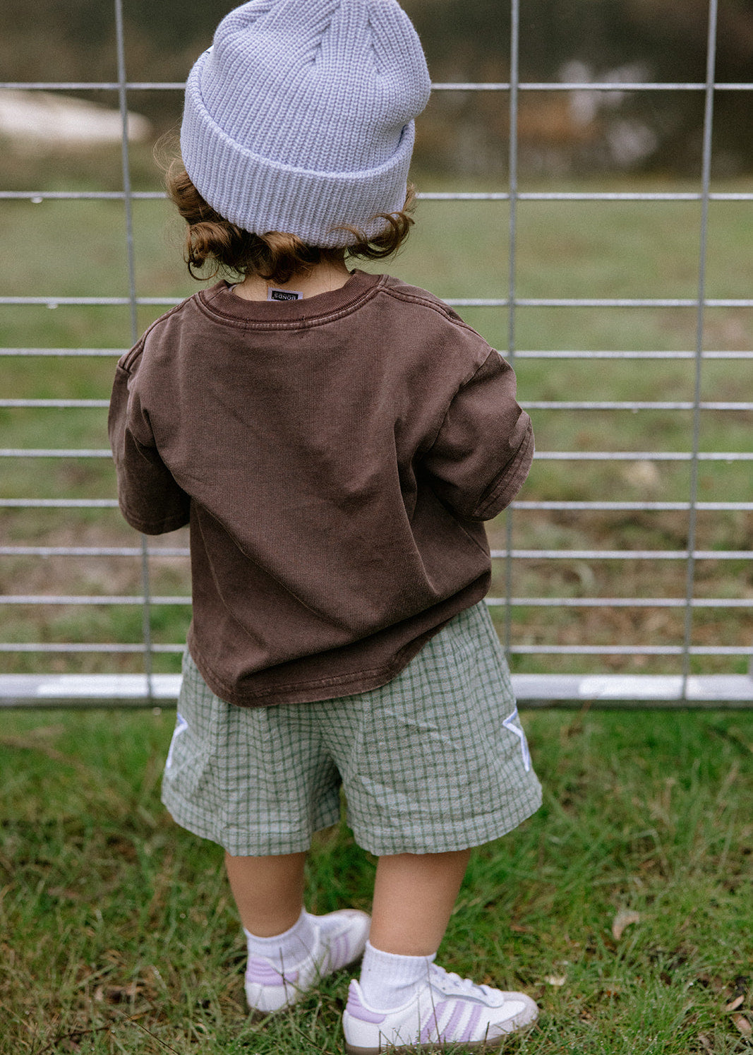 A toddler seen from behind wearing a brown long-sleeve shirt, green and white checkered shorts, a light blue knit beanie, white socks, and white sneakers, standing on grass in front of a wire fence.