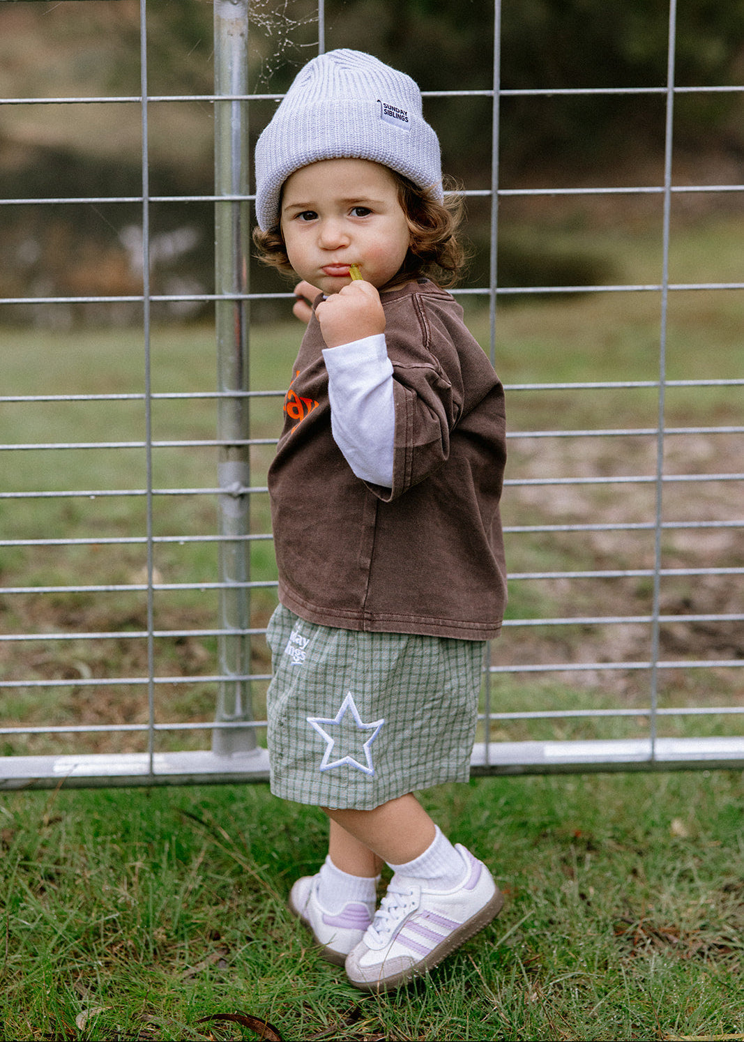 A toddler standing outdoors in front of a wire fence, wearing a light gray knit beanie, a brown corduroy jacket over a white long-sleeve shirt, a green plaid skirt with a white star, white socks, and white sneakers.