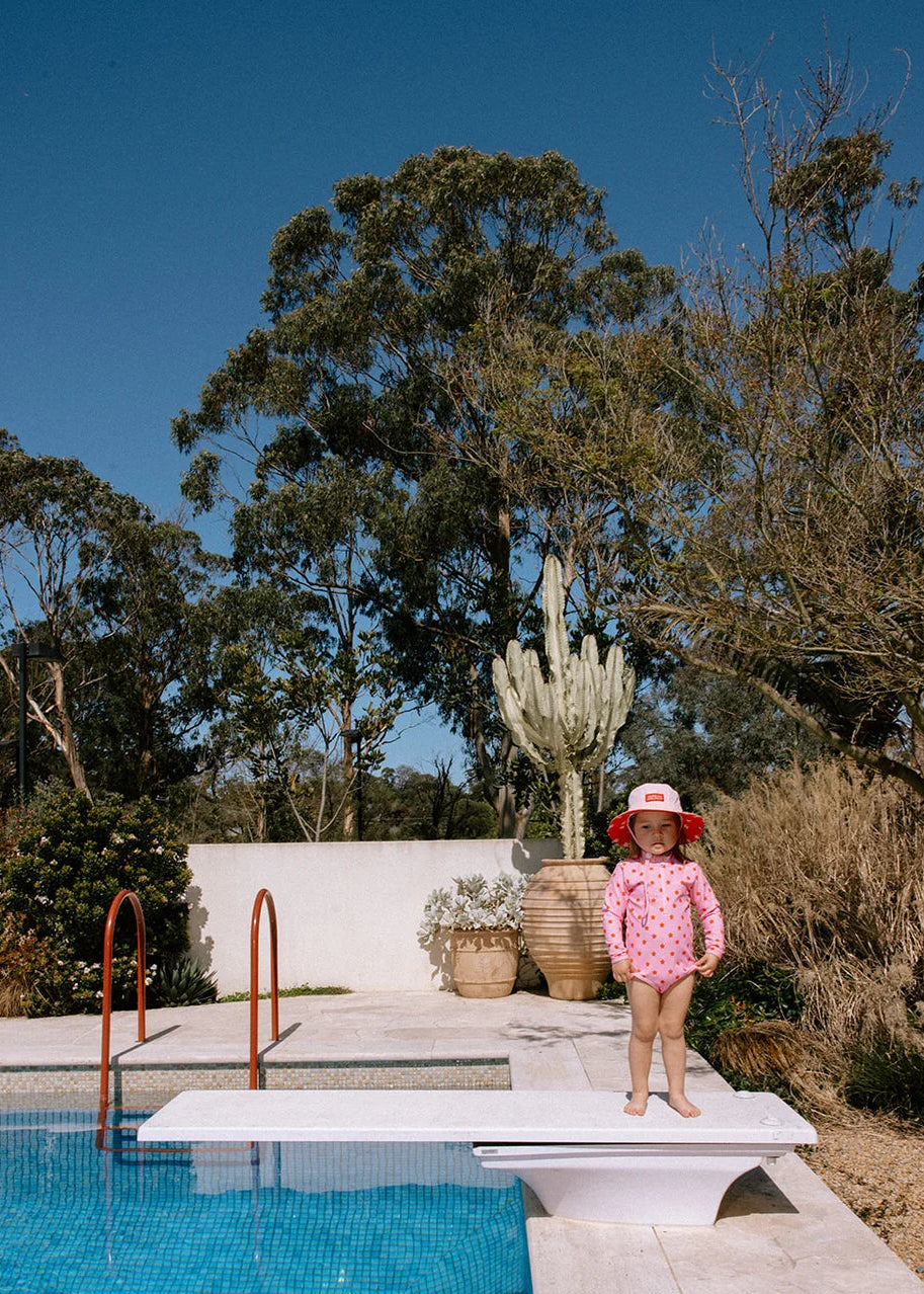 A young child wearing a pink polka-dot swimsuit and a red sun hat stands on a white diving board next to a blue swimming pool, with trees and a white wall in the background.