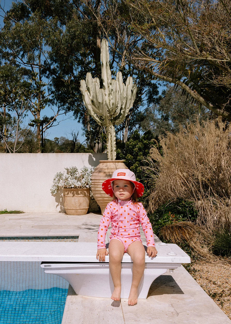 A toddler wearing a pink long-sleeve swimsuit with red polka dots and a matching red sun hat sits on the edge of a swimming pool in an outdoor backyard setting with a cactus and trees in the background.