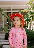 A young child wearing a pink long-sleeve one-piece swimsuit with orange star patterns and a matching red cowboy hat with white stars, standing outdoors in front of a house with green plants.