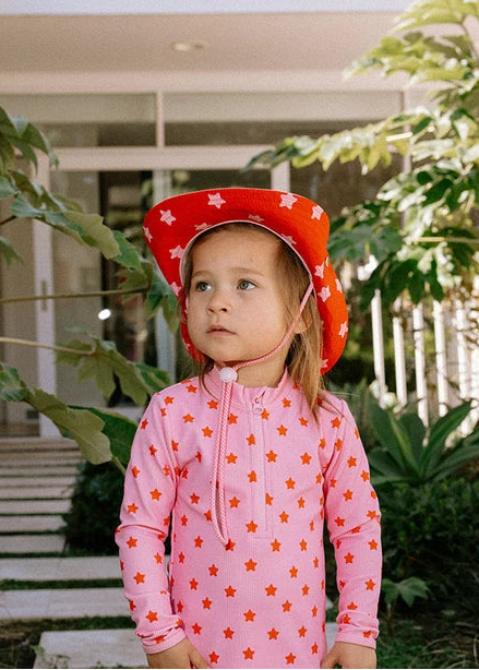 A young child wearing a pink long-sleeve one-piece swimsuit with orange star patterns and a matching red cowboy hat with white stars, standing outdoors in front of a house with green plants.